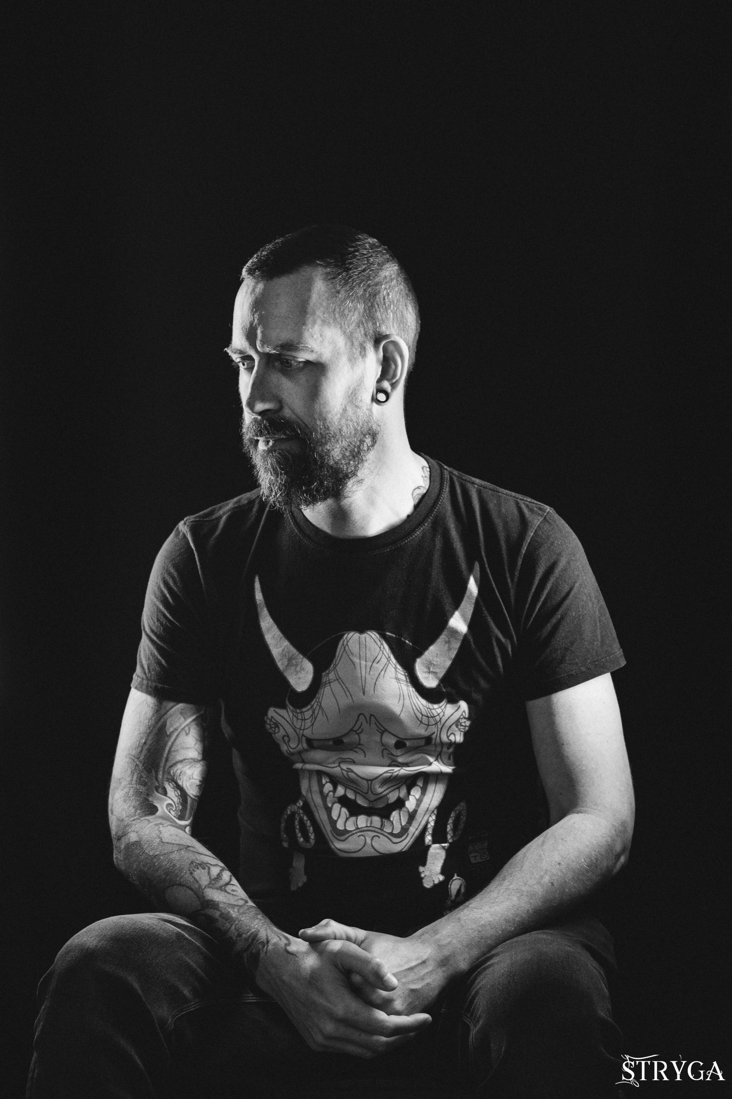 Un homme avec une barbe et des tatouages est assis contre un fond noir, regardant vers le bas. Il porte un t-shirt avec un design de crâne. La photo est en noir et blanc. Le nom 'STRYGA' est écrit en bas à droite.
