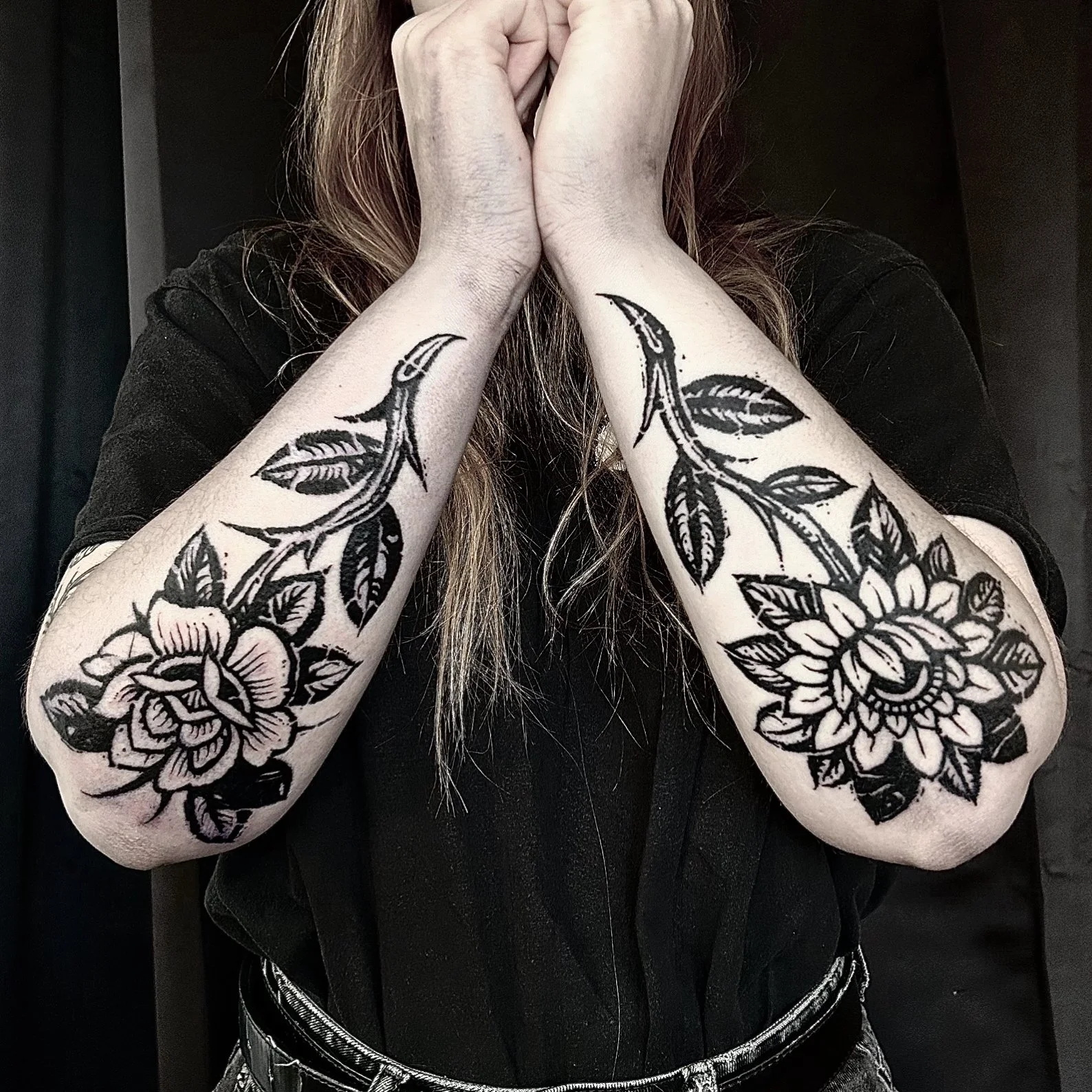 Une personne montre ses bras tatoués avec des motifs floraux noirs, portant un t-shirt noir.