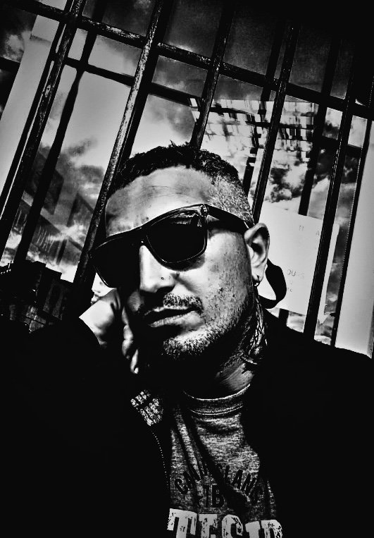 Un homme portant des lunettes de soleil, avec une coiffure en undercut et des dreadlocks, pose devant une grille en métal, dans un environnement urbain.