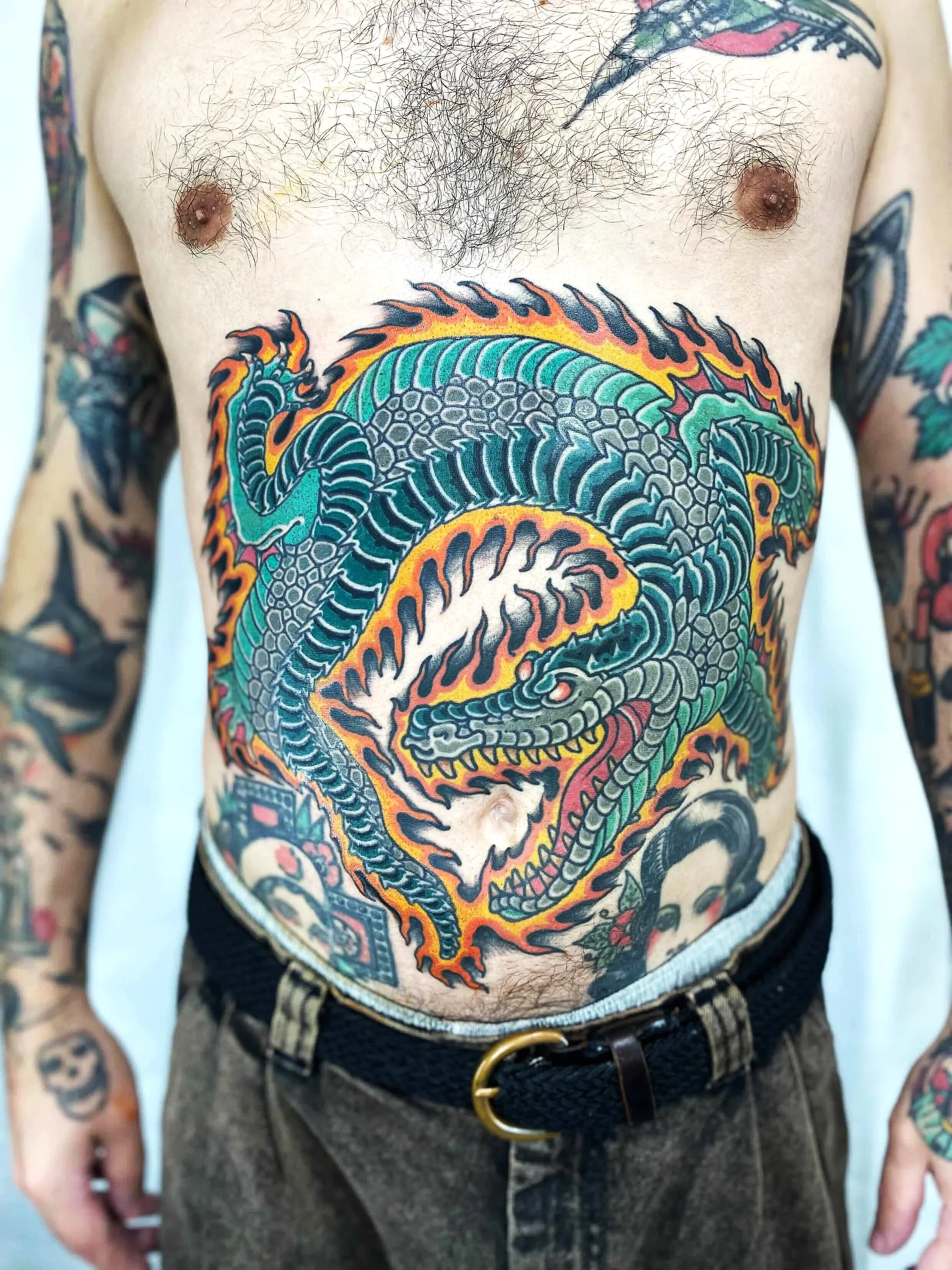 Un torse nu d'un homme avec un tatouage de dragon coloré couvrant une grande partie du torse, avec une boucle de ceinture autour de la taille.