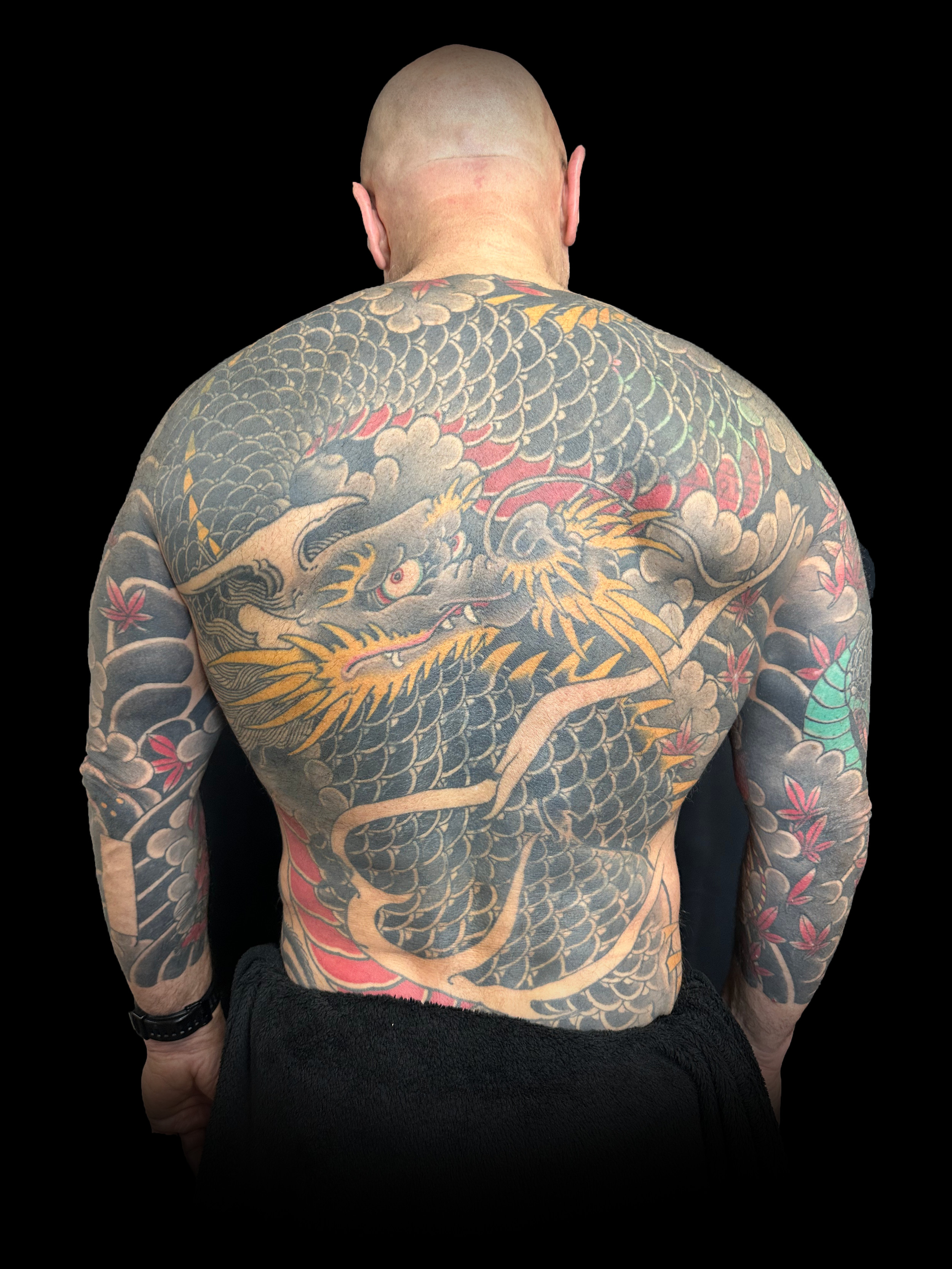 Un homme torse nu avec un dos tatoué d'un grand dragon chinois entouré de nuages et de fleurs, vu de dos, avec la tête inclinée vers l'avant, sur fond noir.