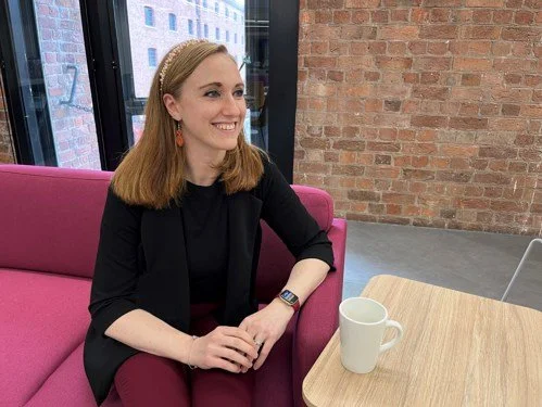 A woman with shoulder-length red hair wearing a black blazer and red pants, sitting on a pink sofa in a modern office space with large windows and exposed brick wall. There is a wooden table with a white mug in front of her.