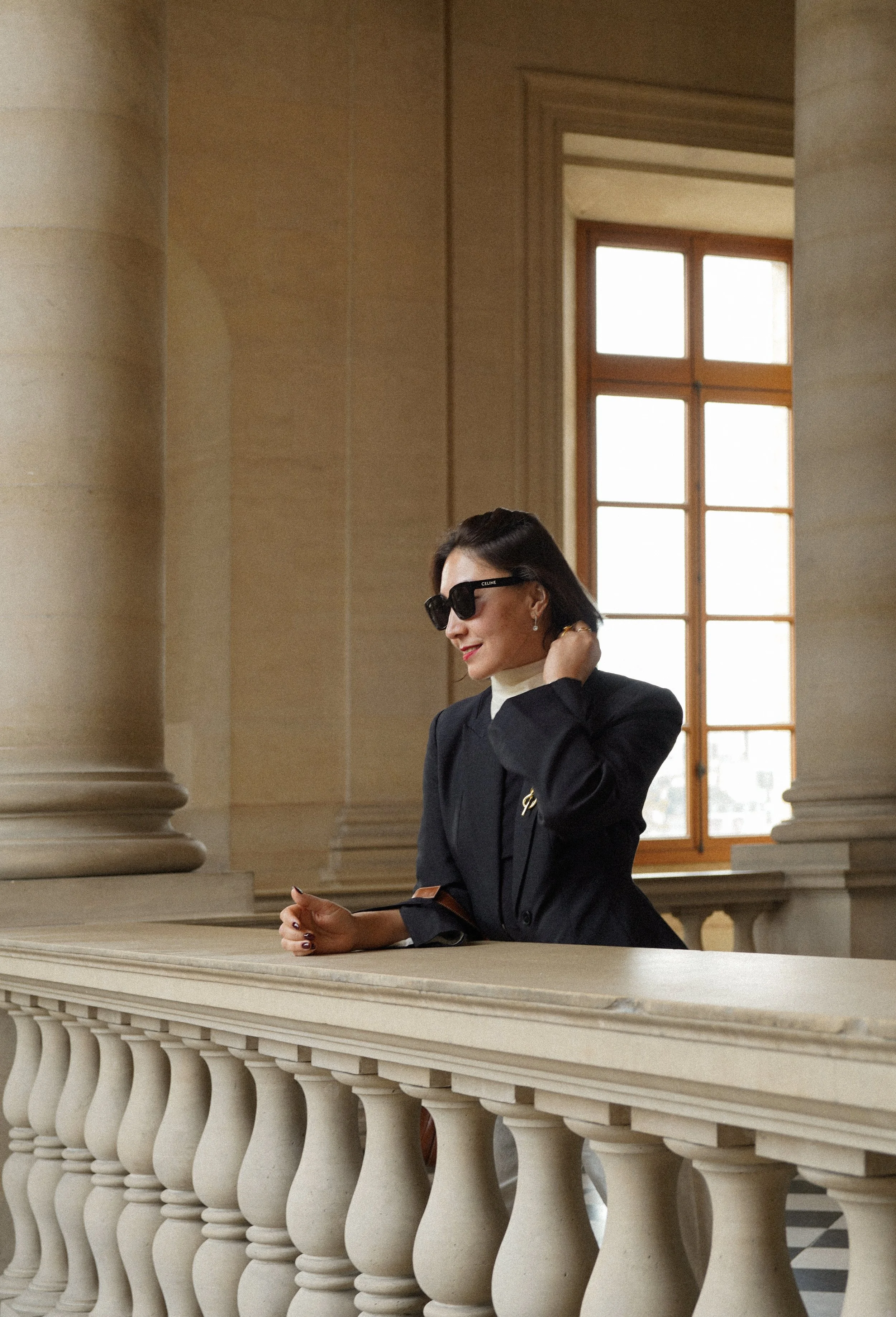 A woman wearing sunglasses and a black blazer standing at a beige stone railing inside a building with large windows.