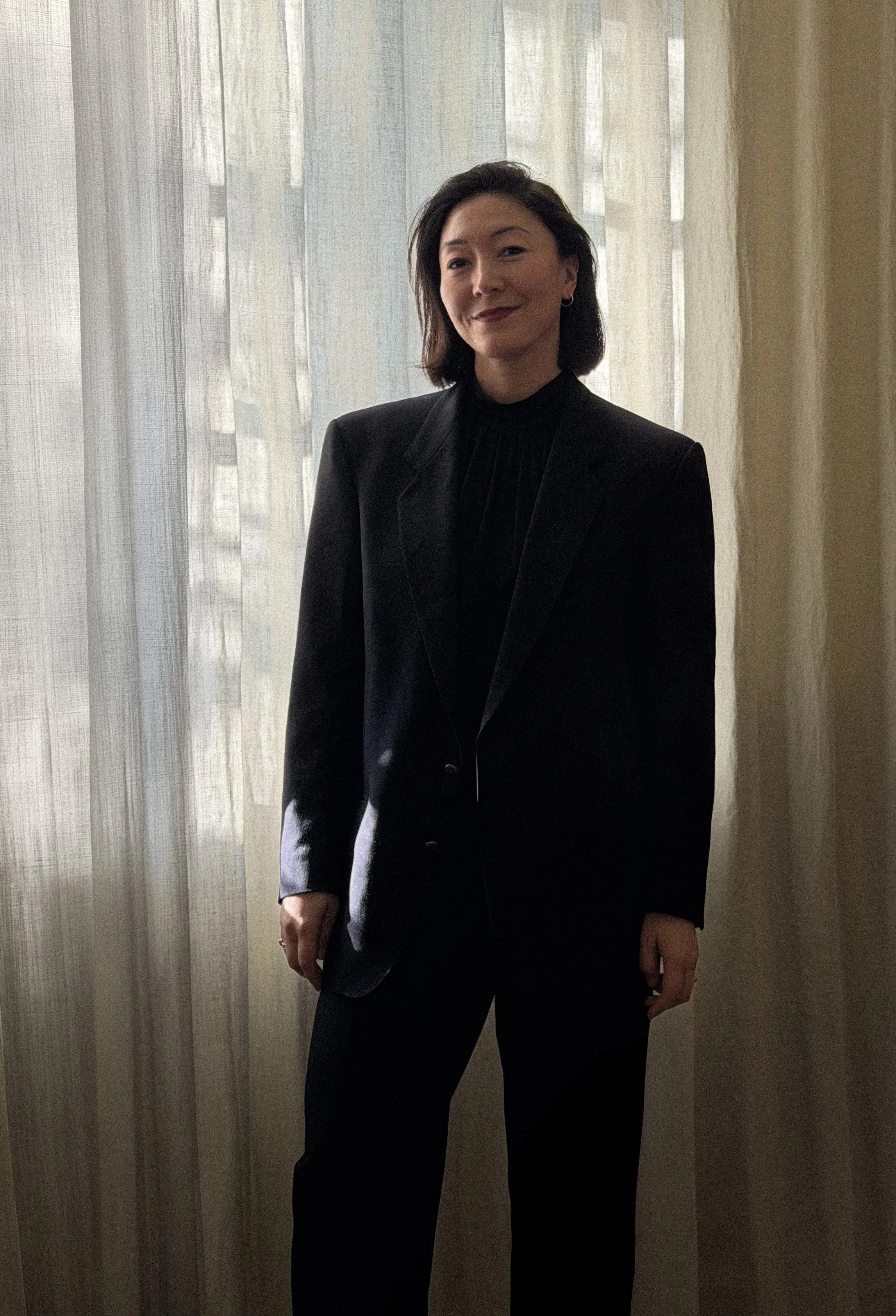 A woman standing indoors in front of beige curtains, wearing a black blazer and black pants, with shoulder-length dark hair and earrings, smiling softly.