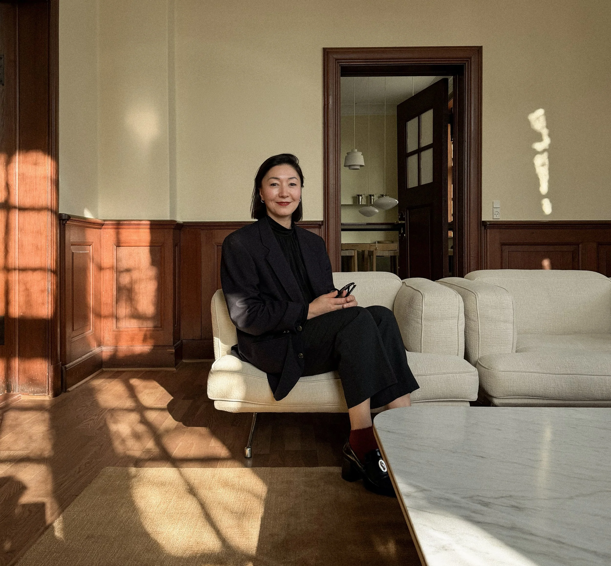 A woman with dark hair sits on a beige sofa in a room with wood-paneled walls and a doorway leading into a kitchen. She is wearing a black blazer and pants, holding a phone, and has a slight smile.
