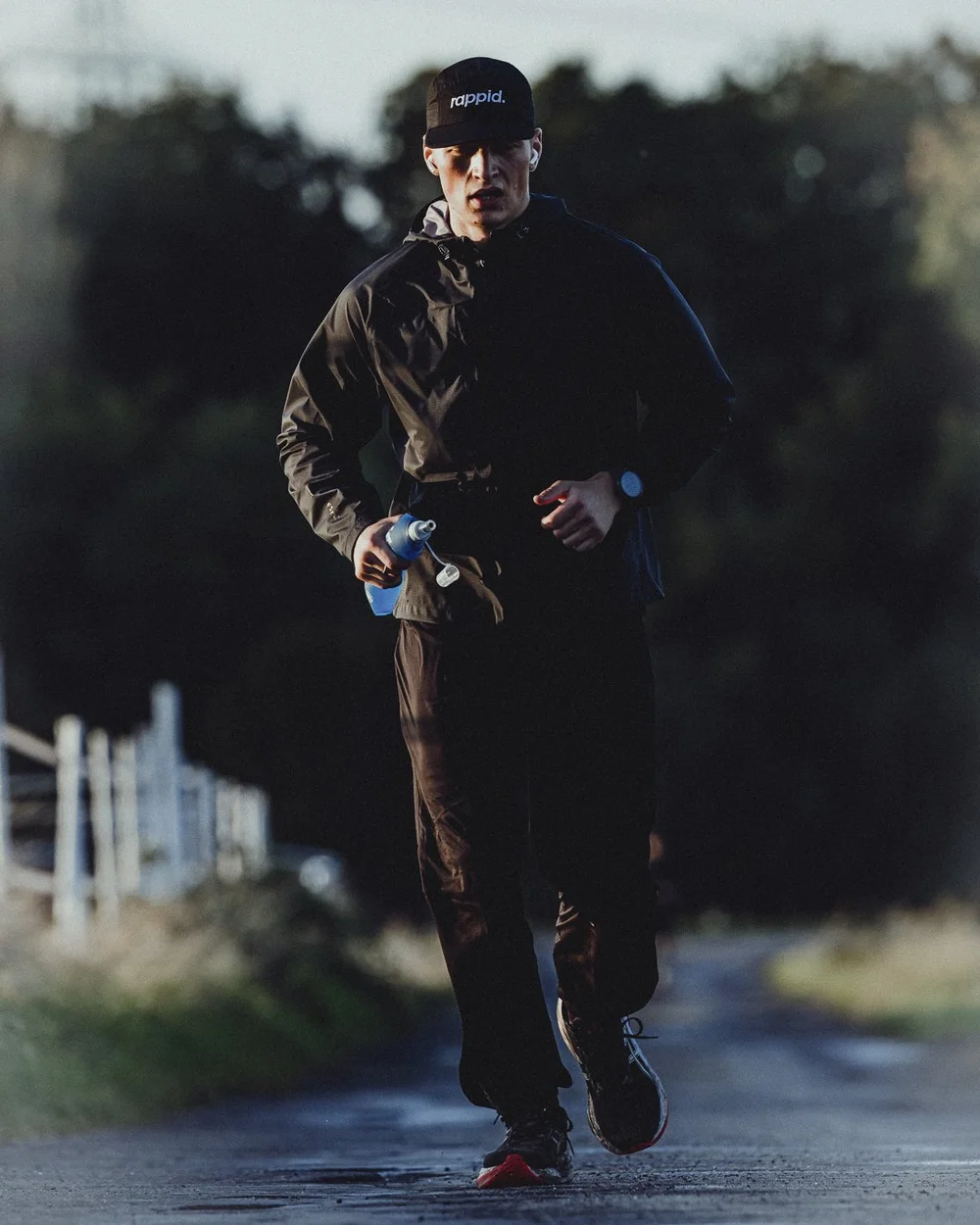 A runner in a black jacket and rappid. cap running back down a wet country road at dawn