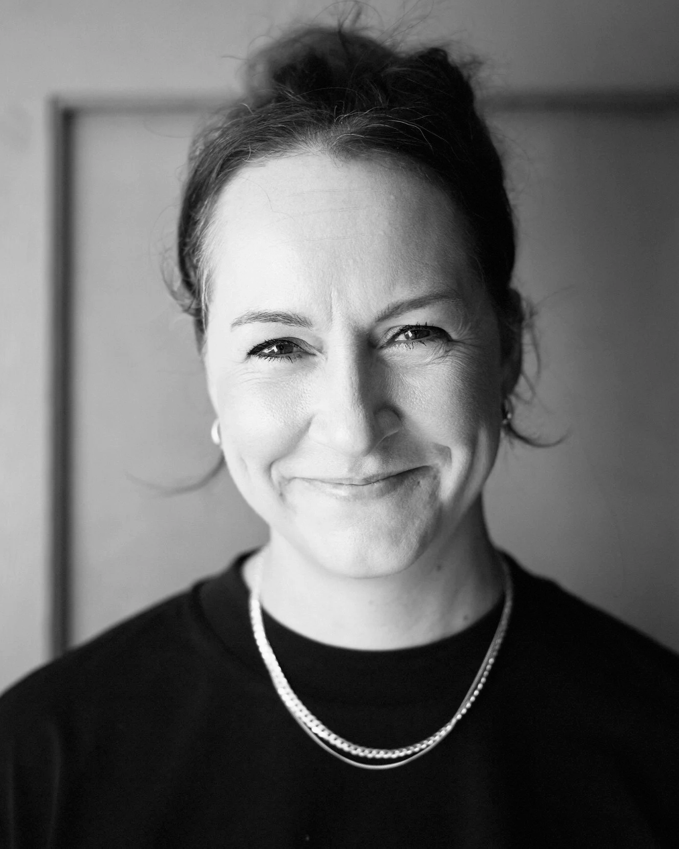 Schwarz-Weiß-Foto einer lächelnden Frau mit dunklen Haaren, die zu einer Seite gestylt sind, trägt eine Halskette und ein dunkles Oberteil.