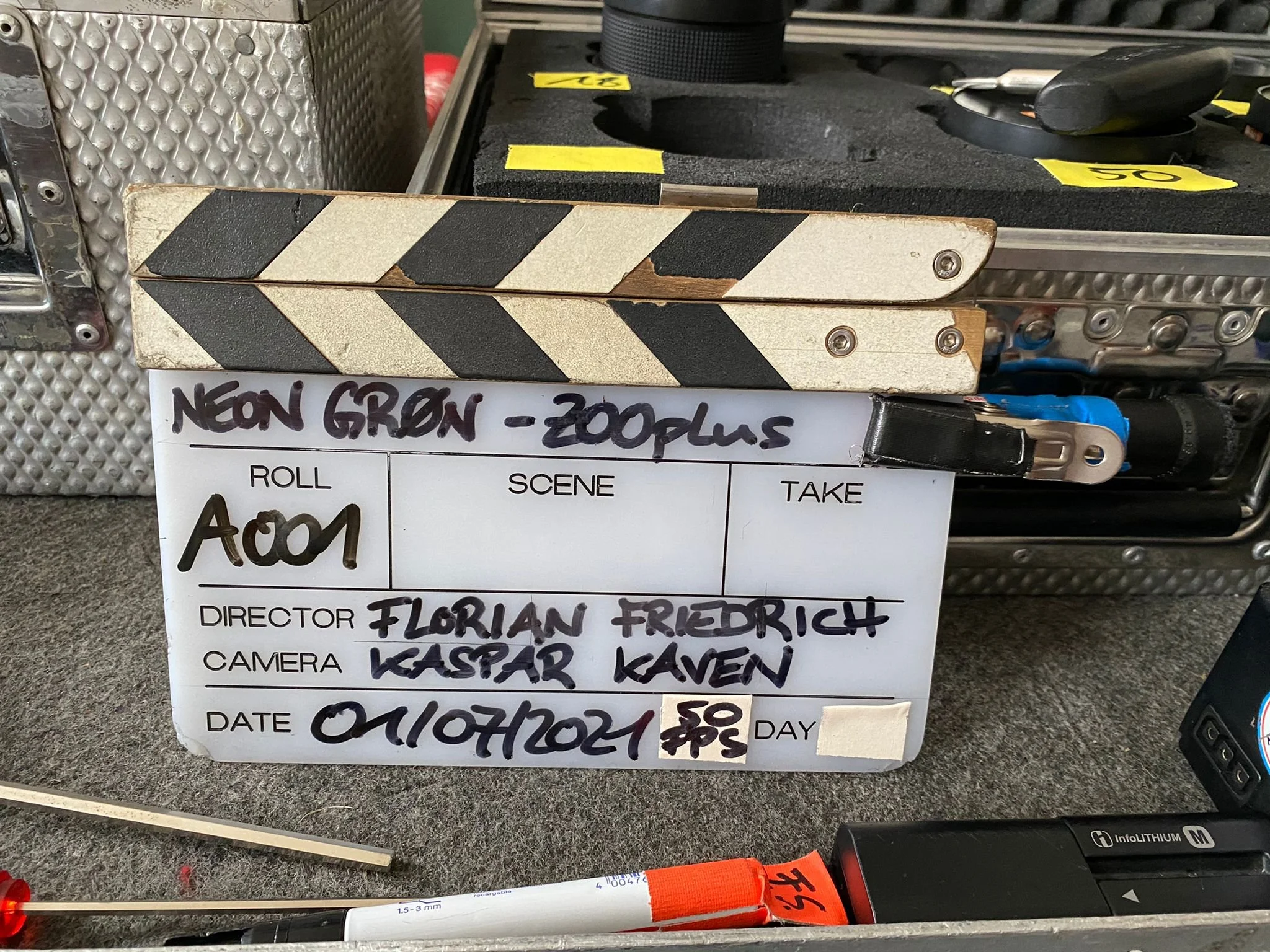 Kladde mit Beschriftung für eine Filmszene, inklusive Regisseur, Kamera, Datum und technischen Angaben, umgeben von Filmausrüstung.