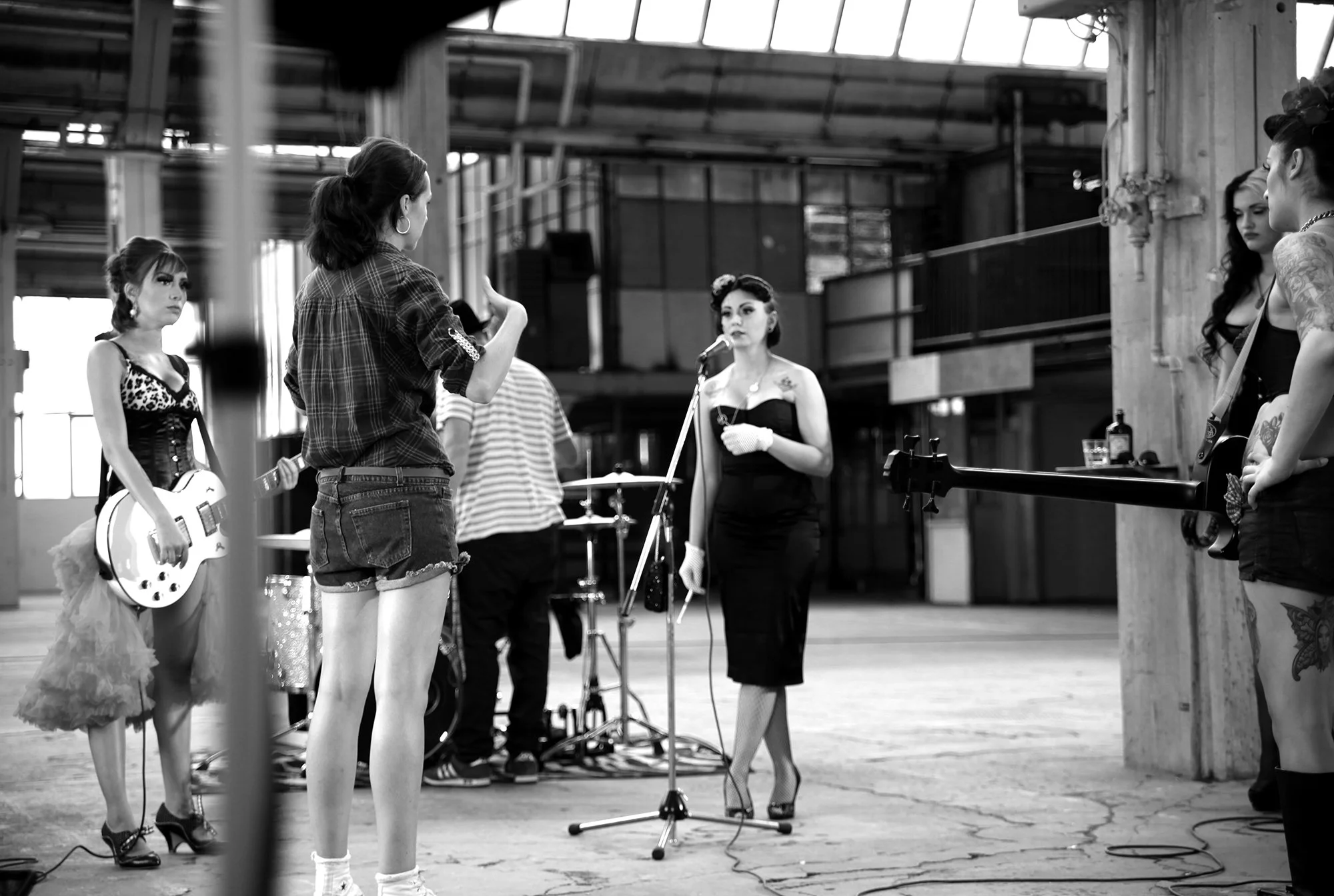 Eine Gruppe von Frauen in einem Industriegebäude, wobei eine Frau mit einem Mikrofon im Mittelpunkt steht, während andere Frauen mit Musikinstrumenten und technischen Geräten im Hintergrund sind.