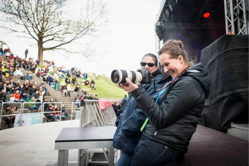 Zwei Frauen auf einer Bühne mit Kamera, während eines Live-Events oder Festivals, mit einem Publikum im Hintergrund.