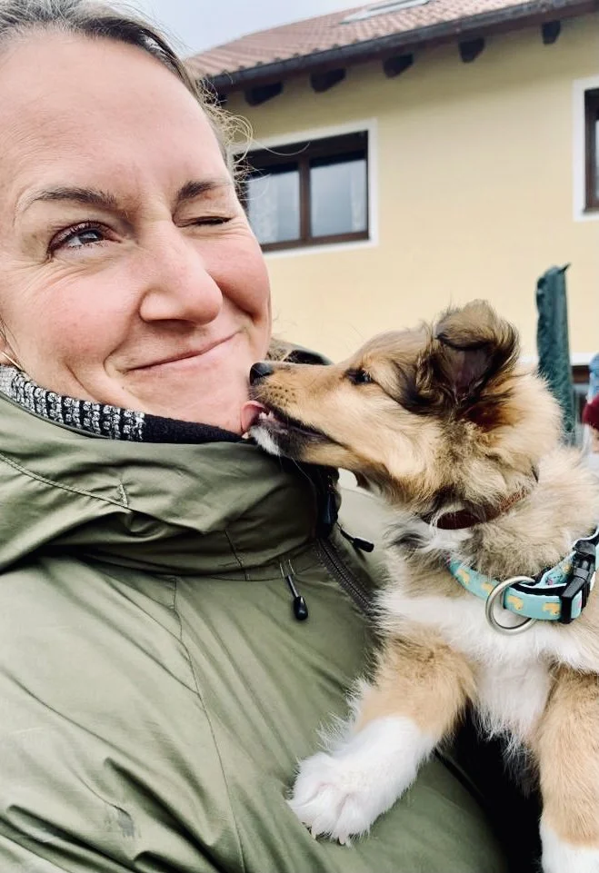 Eine Frau hält einen Welpen, der ihn leckt, vor einem Haus.