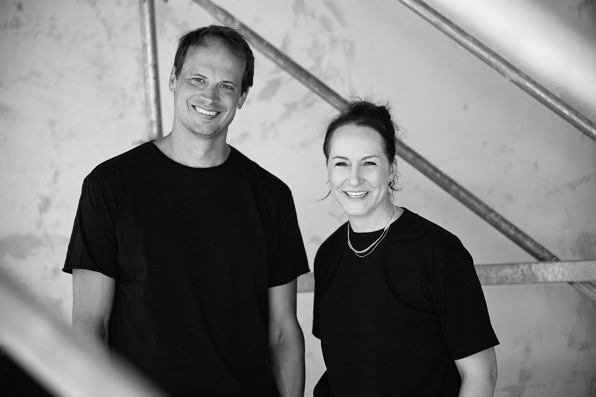 Zwei lachende Menschen, ein Mann und eine Frau, stehen nebeneinander vor einer Wand.