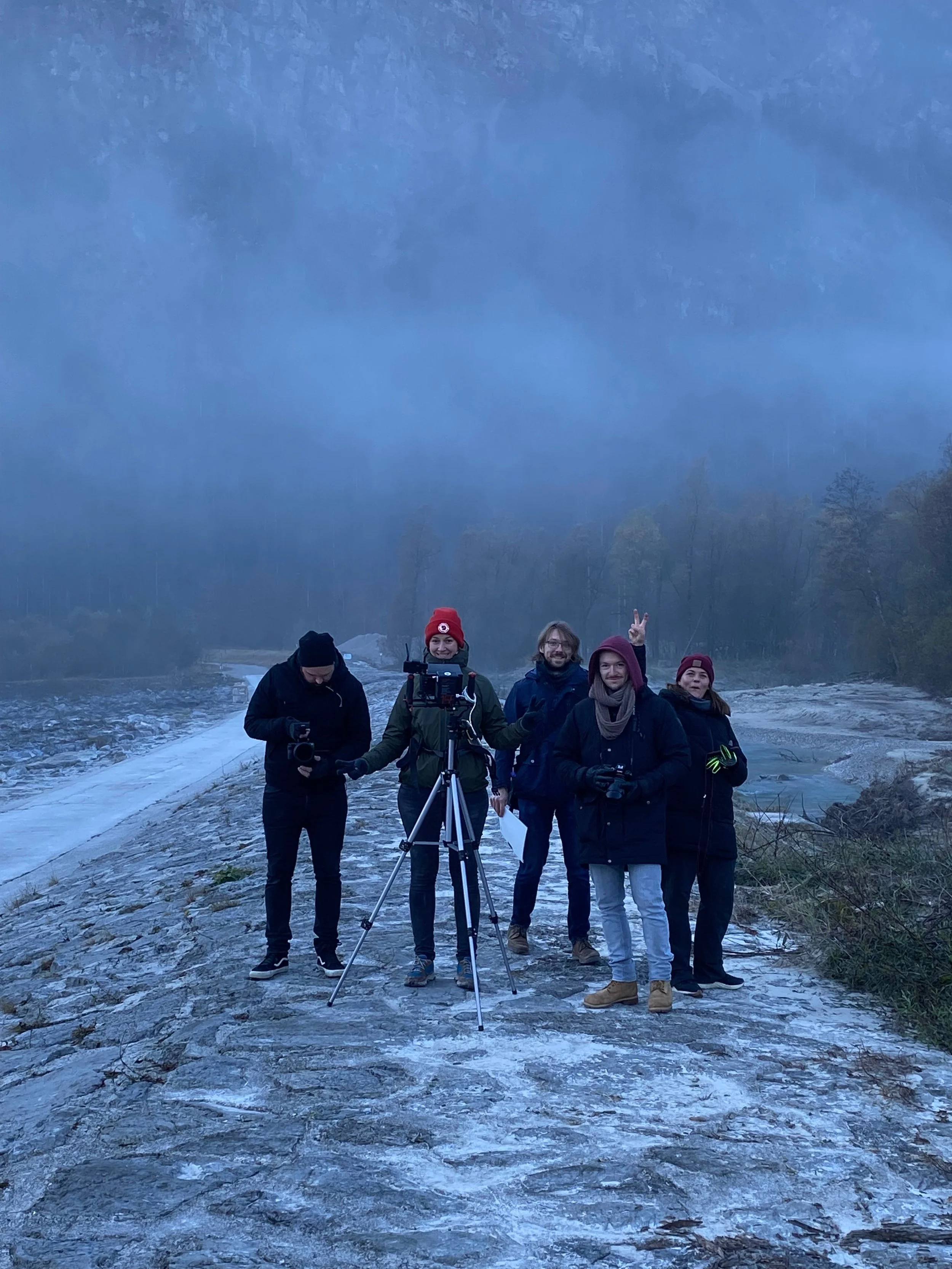 Fünf Personen stehen auf einem felsigen Flussufer im Nebel, einige mit Kameraausrüstung, umgeben von Natur, bei winterlichem Wetter.