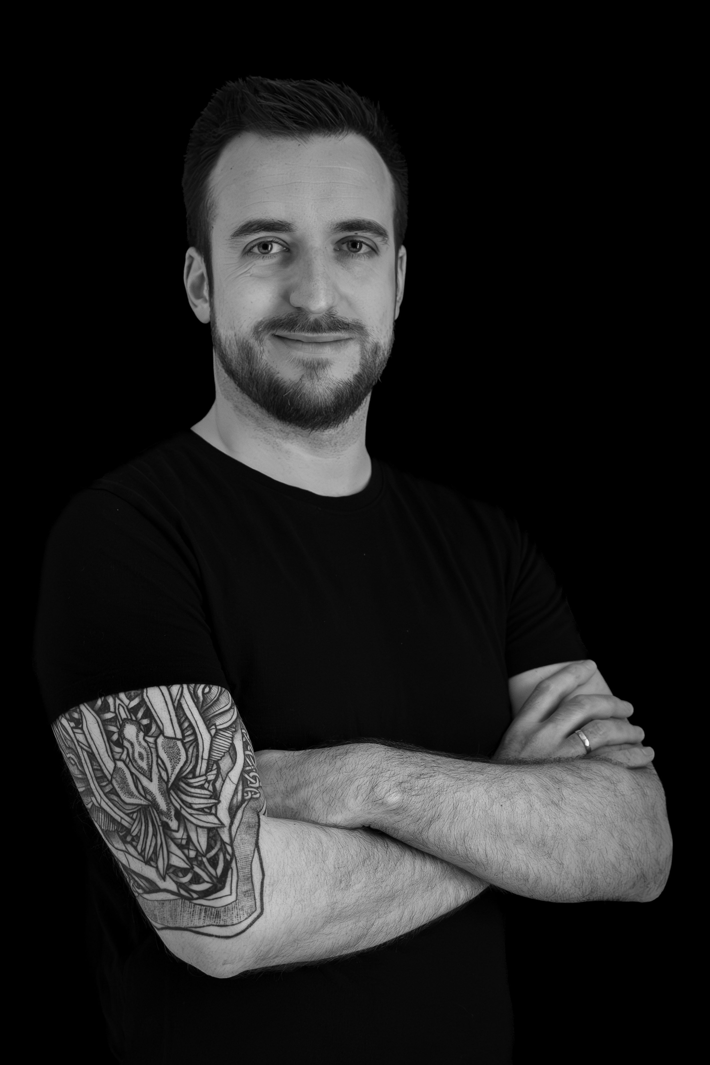 Homme avec barbe, bras croisés, tatouage sur le bras gauche, portant un t-shirt noir, souriant, fond noir.