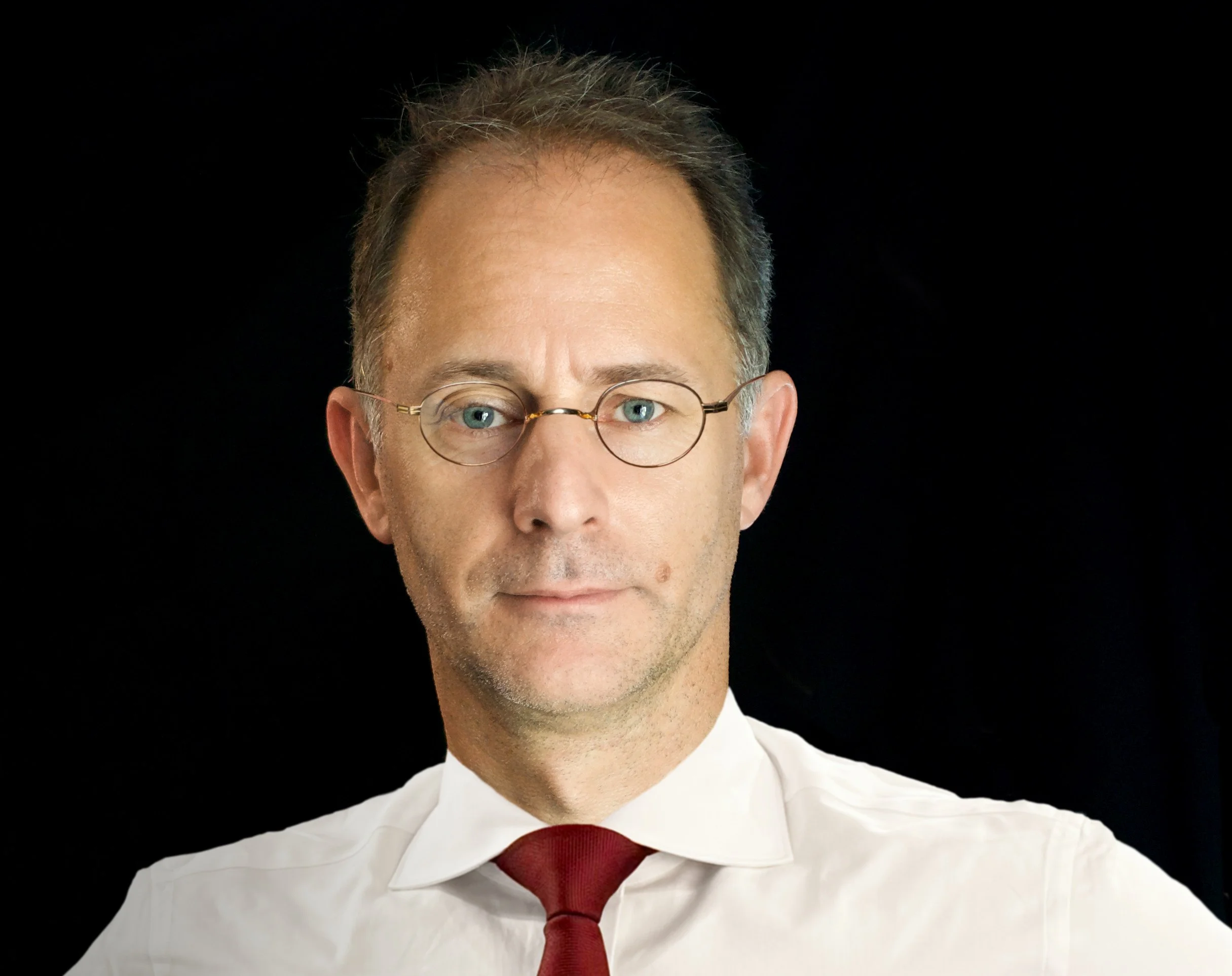 Gedeeltelijke portretfoto van een man met grijs haar, ronde brillen, gekleed in een wit overhemd en een rode tie, tegen een zwarte achtergrond.