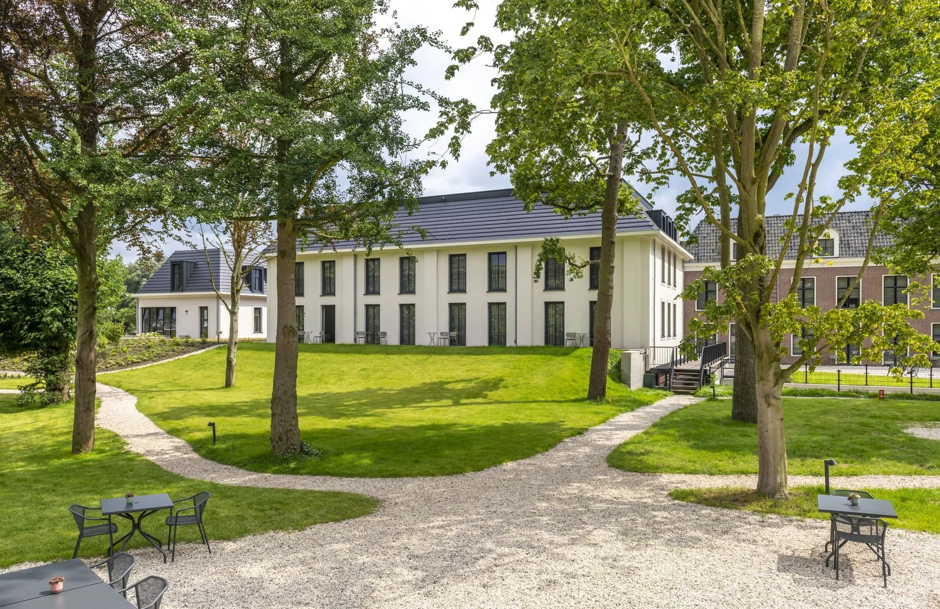 Landschap met gras, wandelpaden, bomen en moderne witte gebouwen met grote ramen in een groene tuinomgeving.