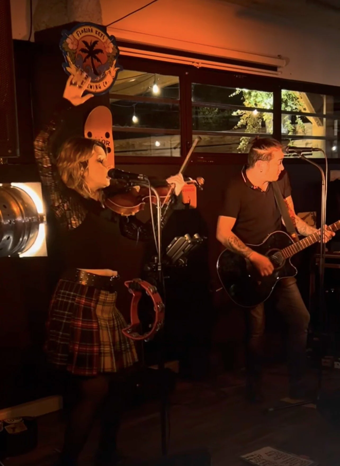 Deux musiciens jouent dans un bar, une femme avec un violon et un tambourin, un homme avec une guitare, dans une ambiance chaleureuse et intime.