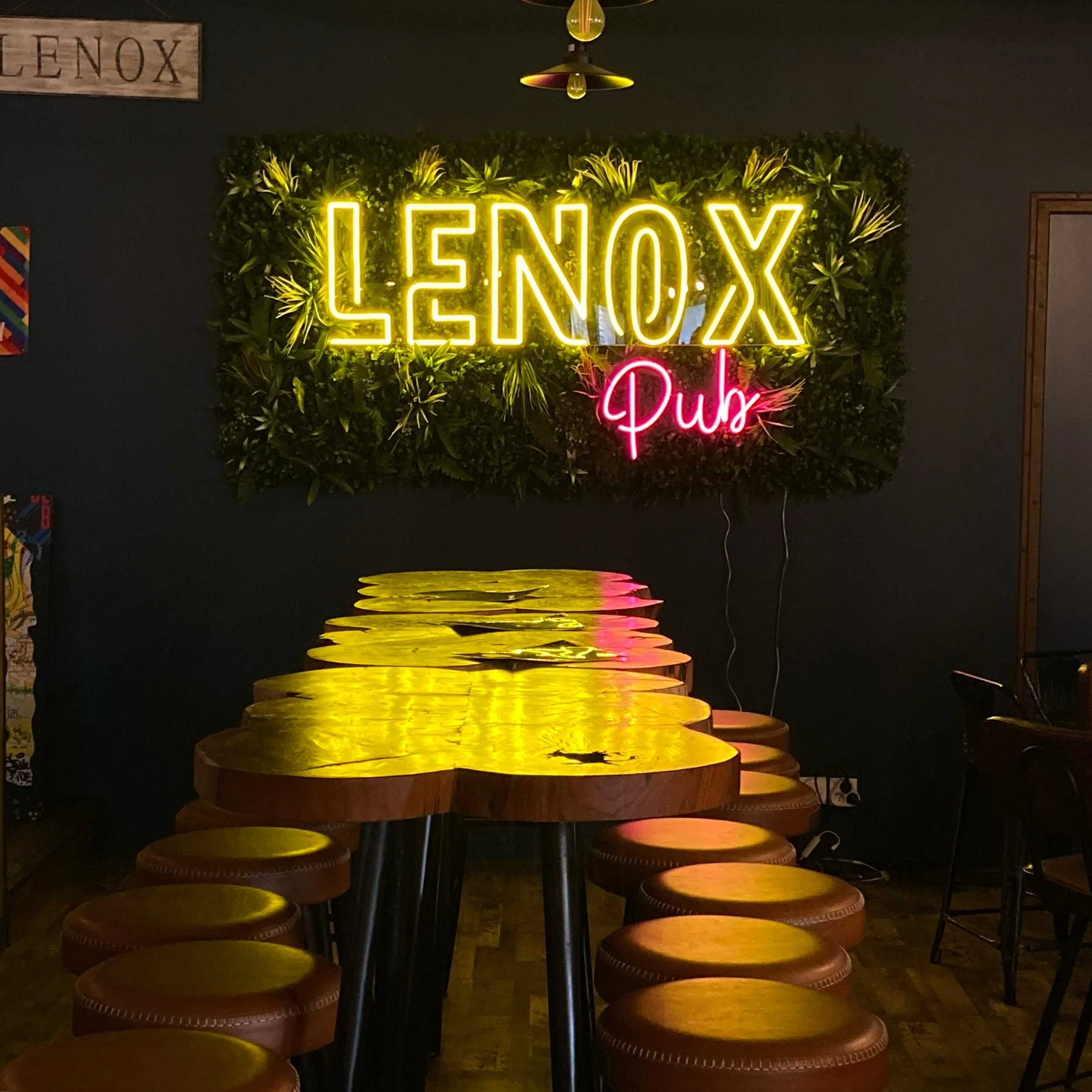 Intérieur d'un bar avec un mur de plantes, un panneau lumineux « LENOX Pub » en néon jaune et rose, et une table en bois avec des tabourets en leather marron.