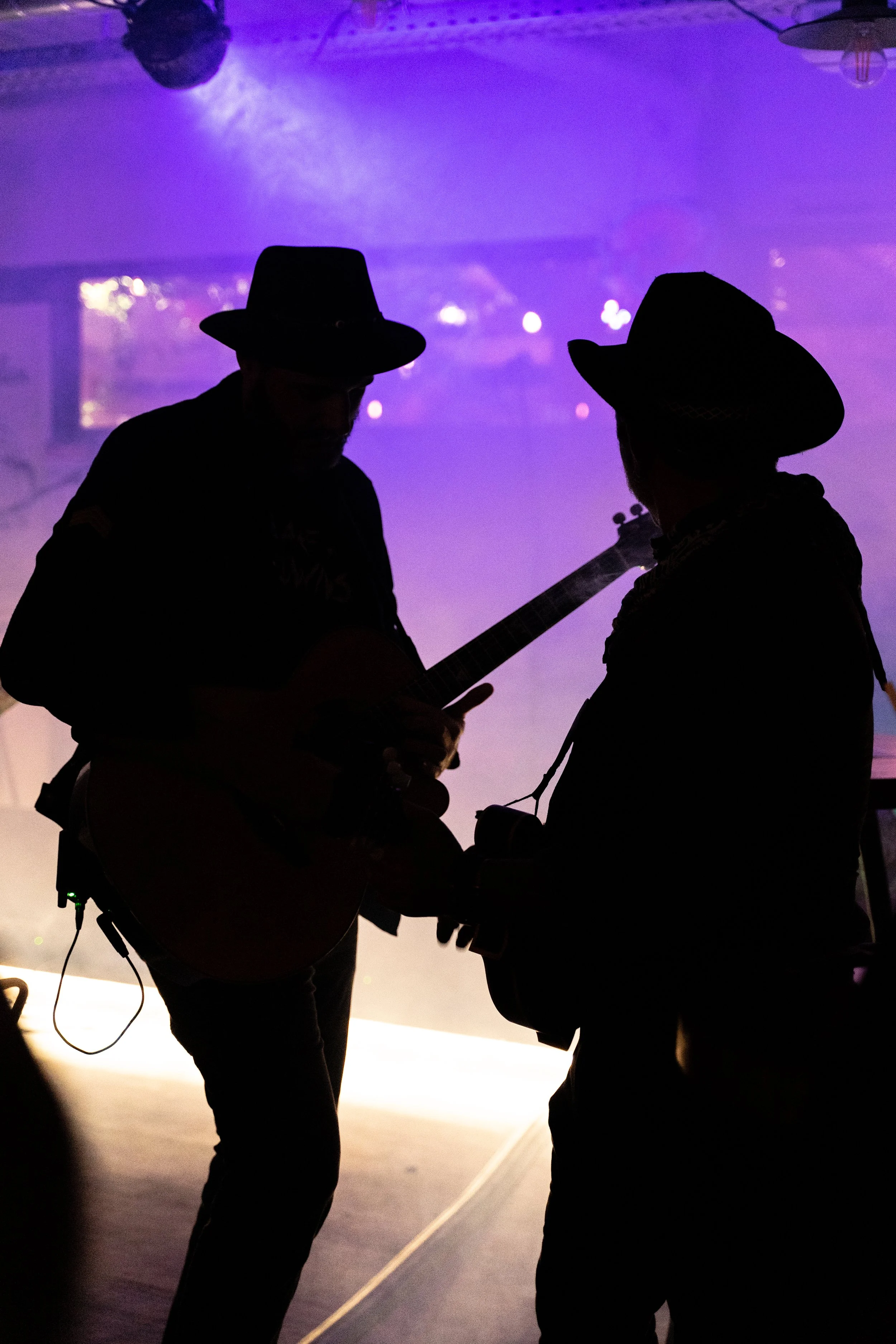 Deux personnes en silhouette portant des chapeaux, jouant de la guitare dans une ambiance sombre avec un éclairage violet en arrière-plan.