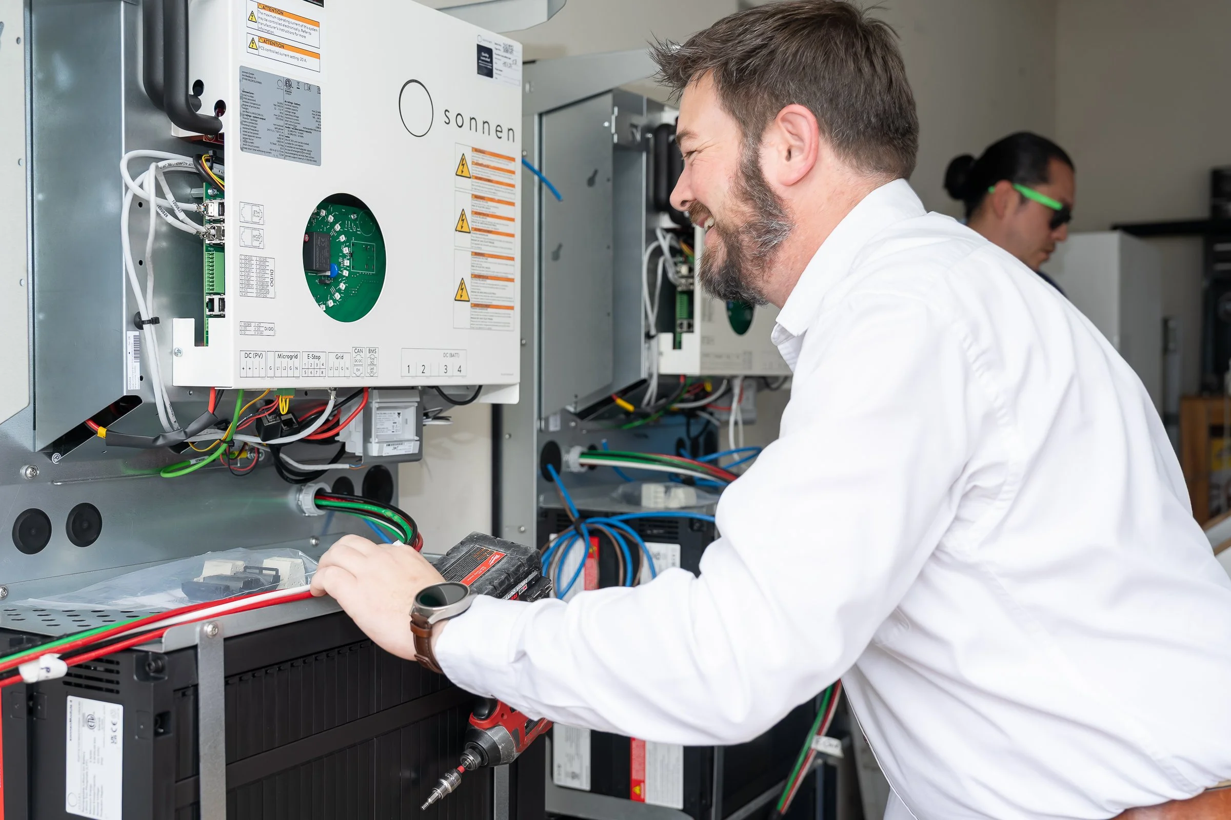 A technician working on electrical equipment, smiling, while installing a battery for a home owner to become self reliant.