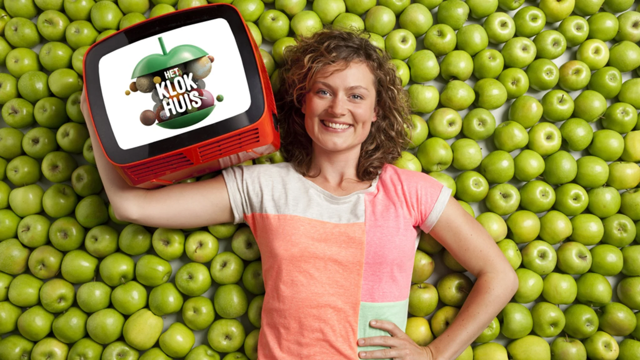 A smiling woman with curly hair lying on a bed of green apples, holding an orange toy box with a logo that says 'Het Klokhuis' and an image of an apple with a hammock.