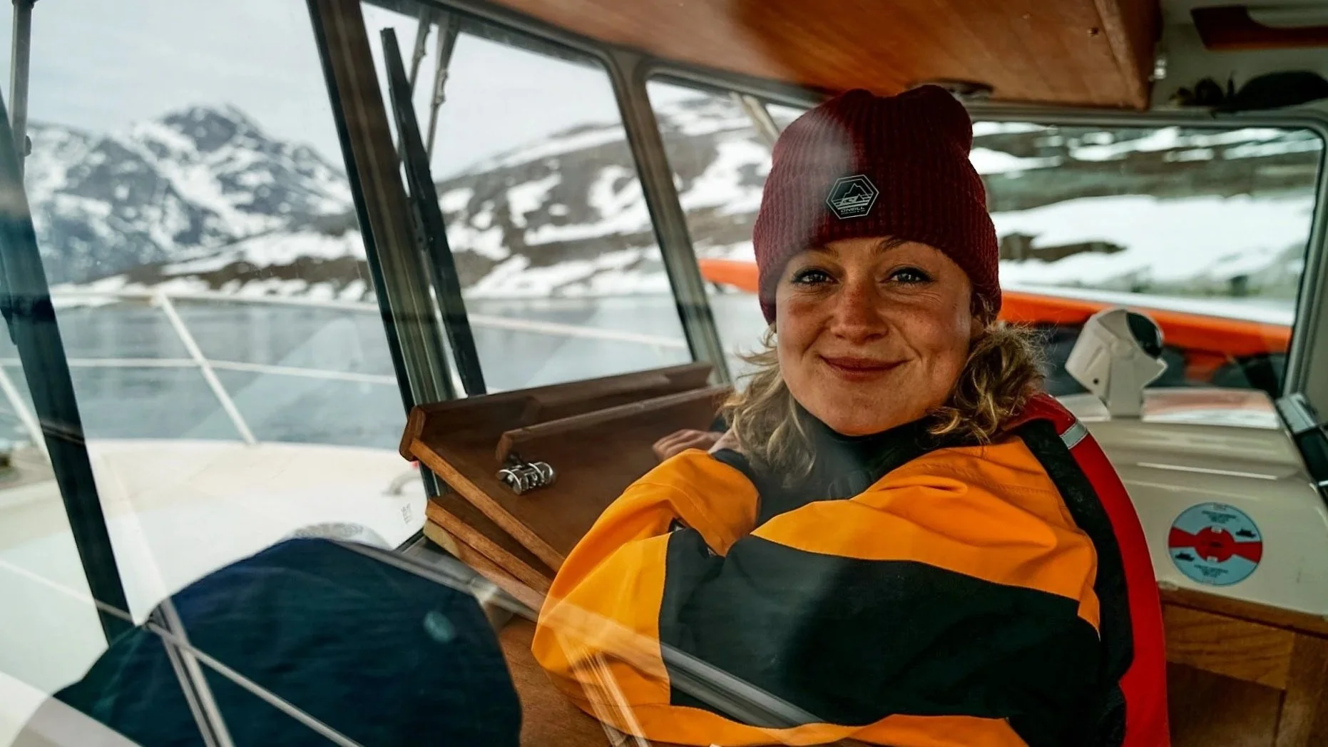 A woman in a red beanie and orange rain jacket sitting inside a boat with a snowy mountain landscape outside the window.