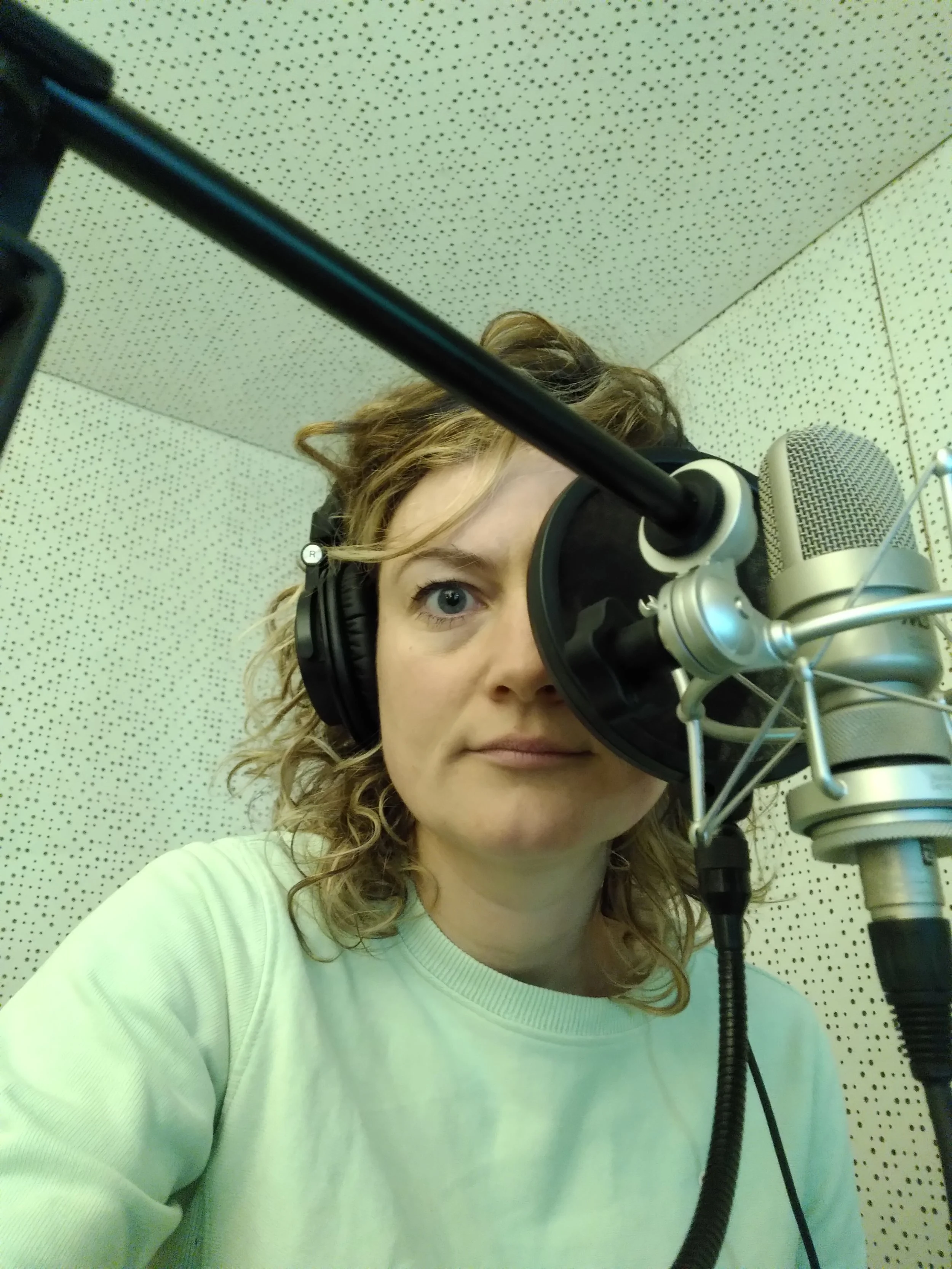 A woman with curly blonde hair wearing headphones in a recording studio, looking at the microphone in front of her.