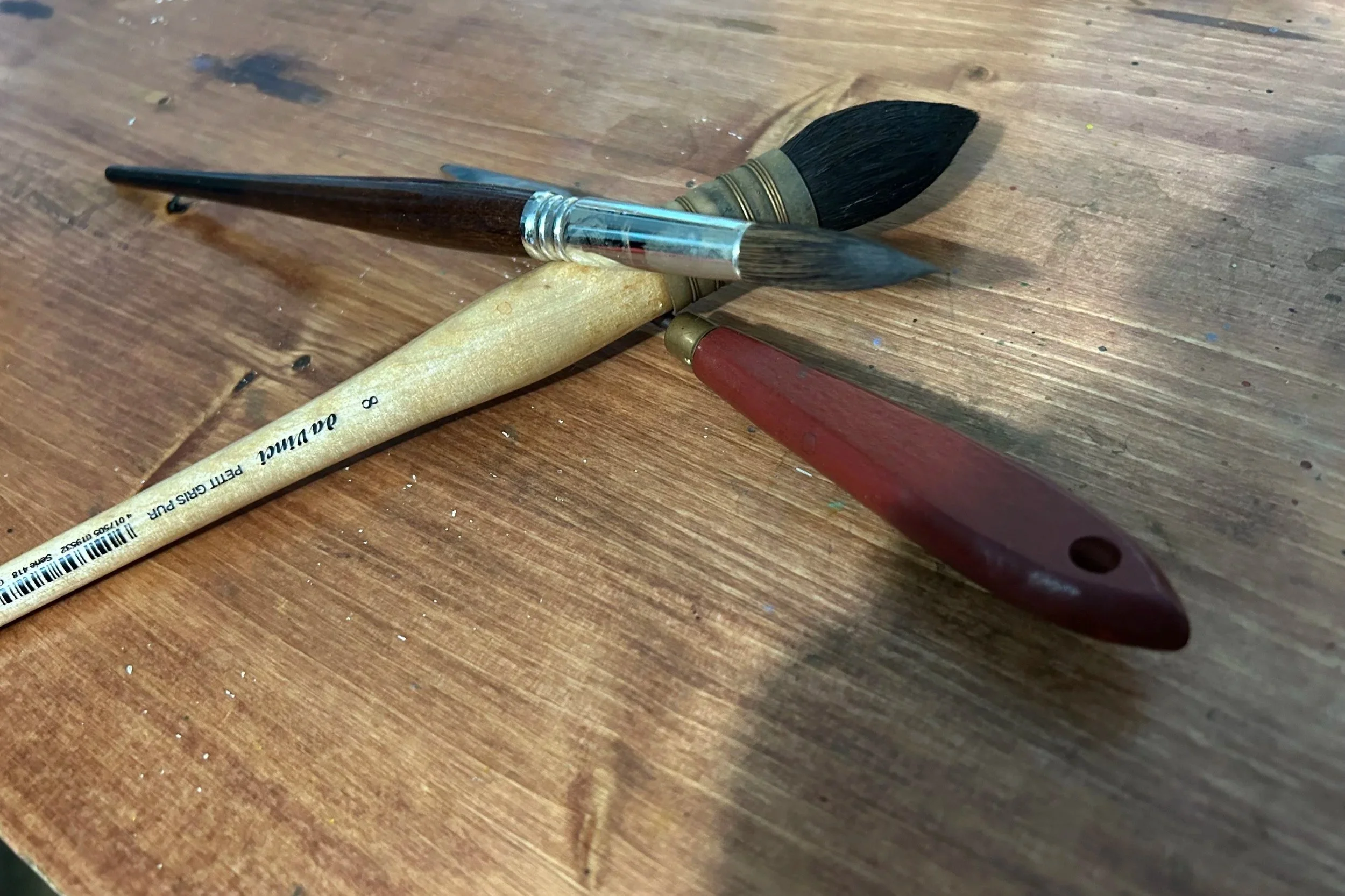 Three paintbrushes on a wooden surface, one with a light-colored handle, one with a dark brown handle, and one with a red handle. The brushes are crossed and oriented diagonally.