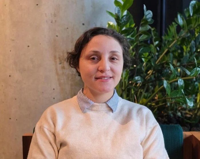 A woman sitting indoors with a concrete wall and green plants in the background, wearing a beige sweatshirt over a collared shirt.