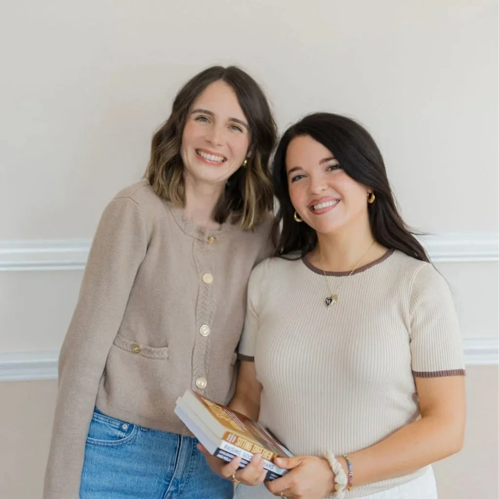 Two women smiling, standing close together indoors against a plain off-white wall. One woman with shoulder-length brown hair in a beige sweater, and the other with long dark hair wearing a white t-shirt with brown trim, holding a book.