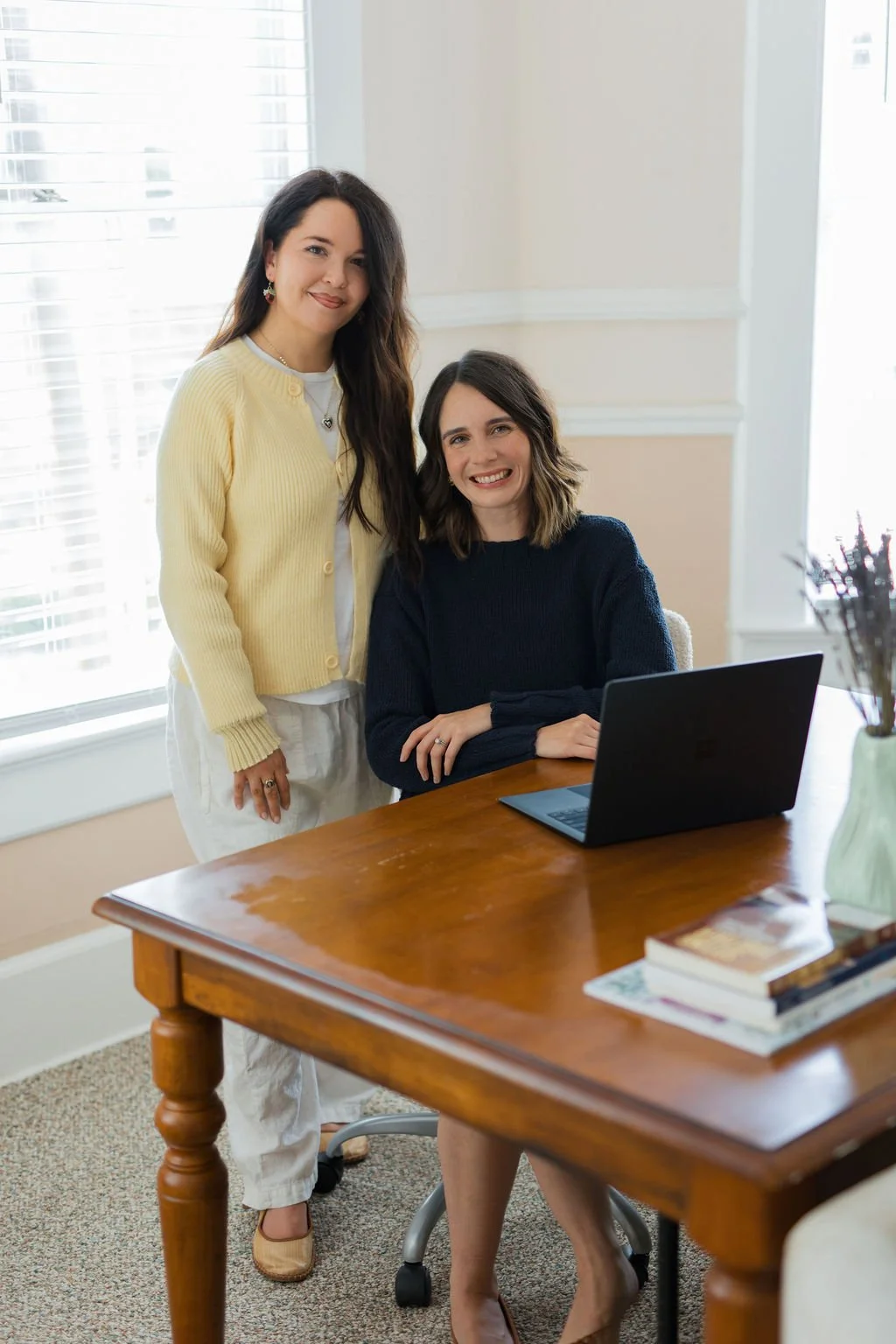 Two women in an office, one standing and one sitting at a desk with a laptop, both smiling.