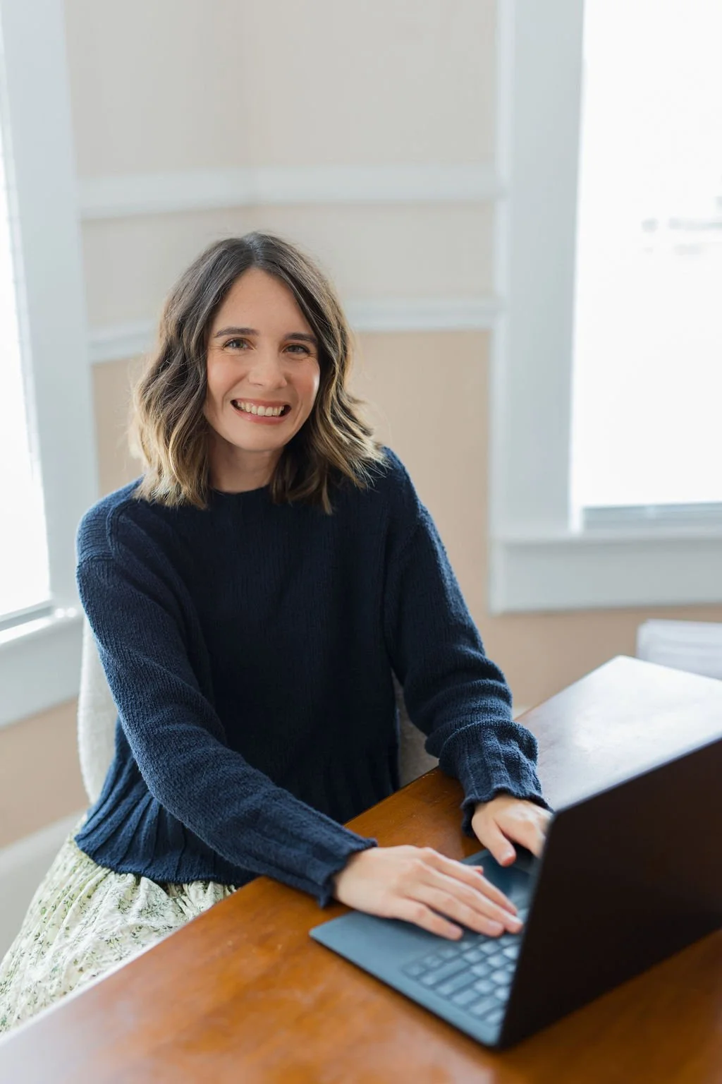 A woman with shoulder-length wavy hair wearing a dark blue sweater, sitting at a wooden desk with a laptop in front of her, smiling at the camera in a well-lit room with large windows.
