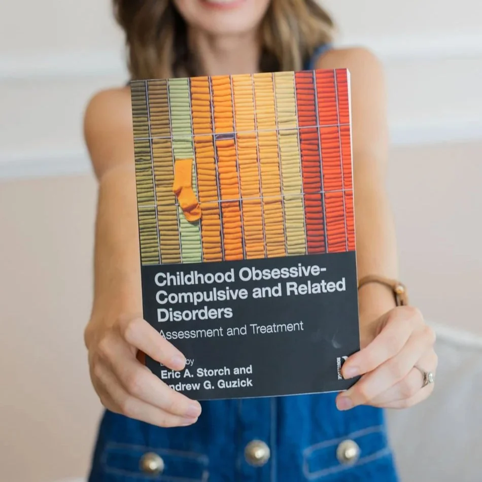 A woman holding a book titled "Childhood Obsessive-Compulsive and Related Disorders: Assessment and Treatment" in front of her face. The cover features a colorful illustration of toothbrushes.