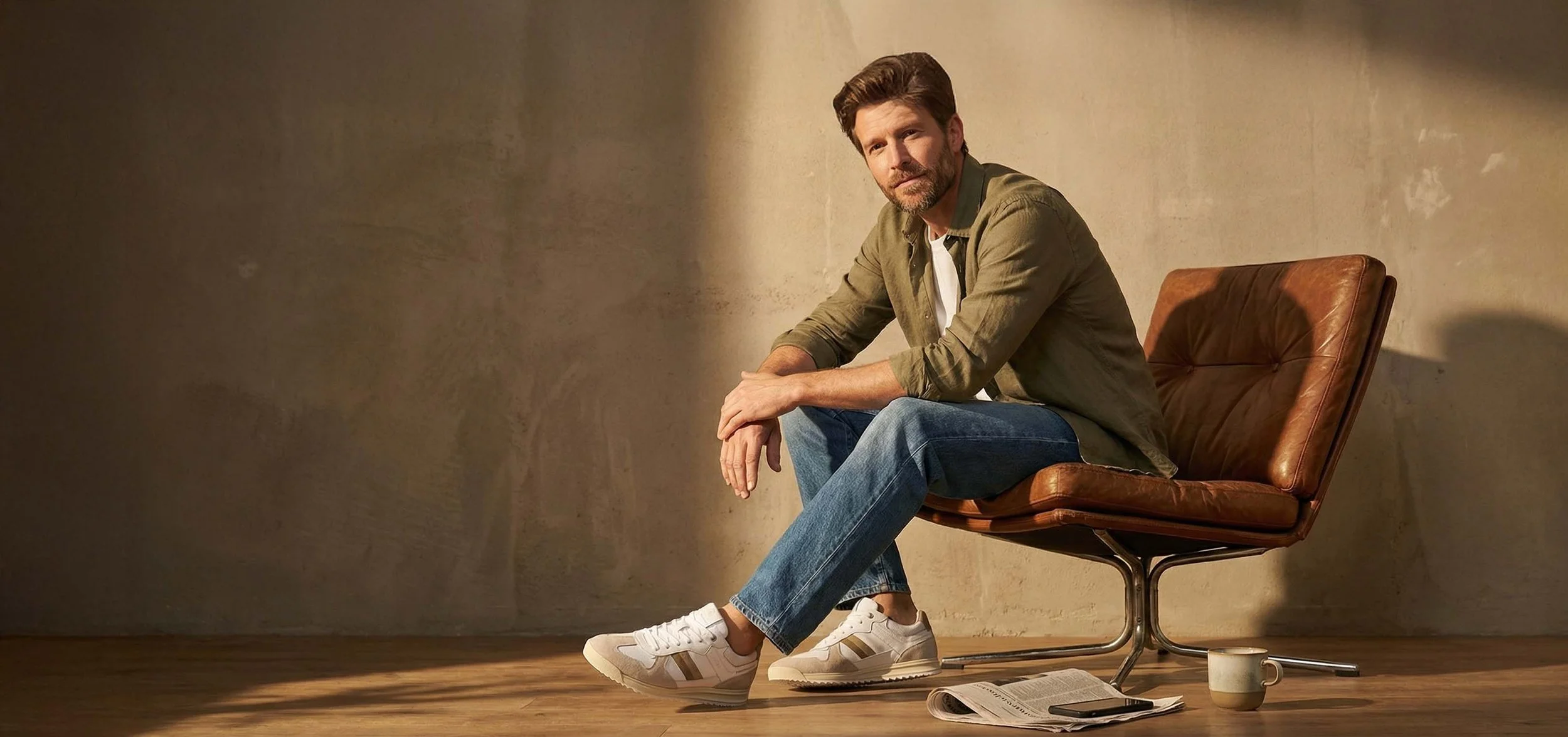 A man with brown hair and a beard sitting on a brown leather chair in front of a textured wall, wearing a green shirt, blue jeans, and white sneakers, with a newspaper, a smartphone, and a mug on the wooden floor.