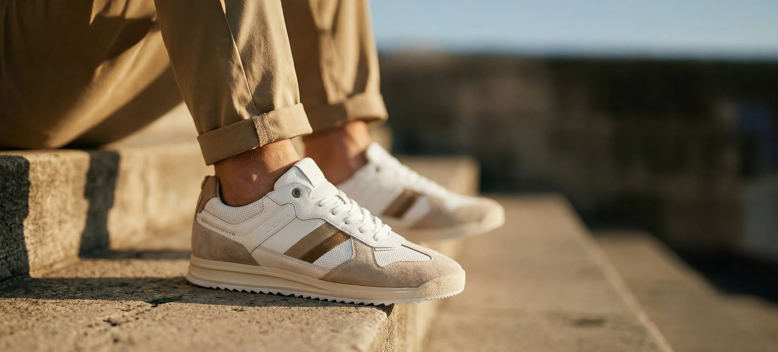 Close-up of a person sitting on outdoor steps, wearing white sneakers with beige and brown accents and tan pants with rolled-up cuffs.