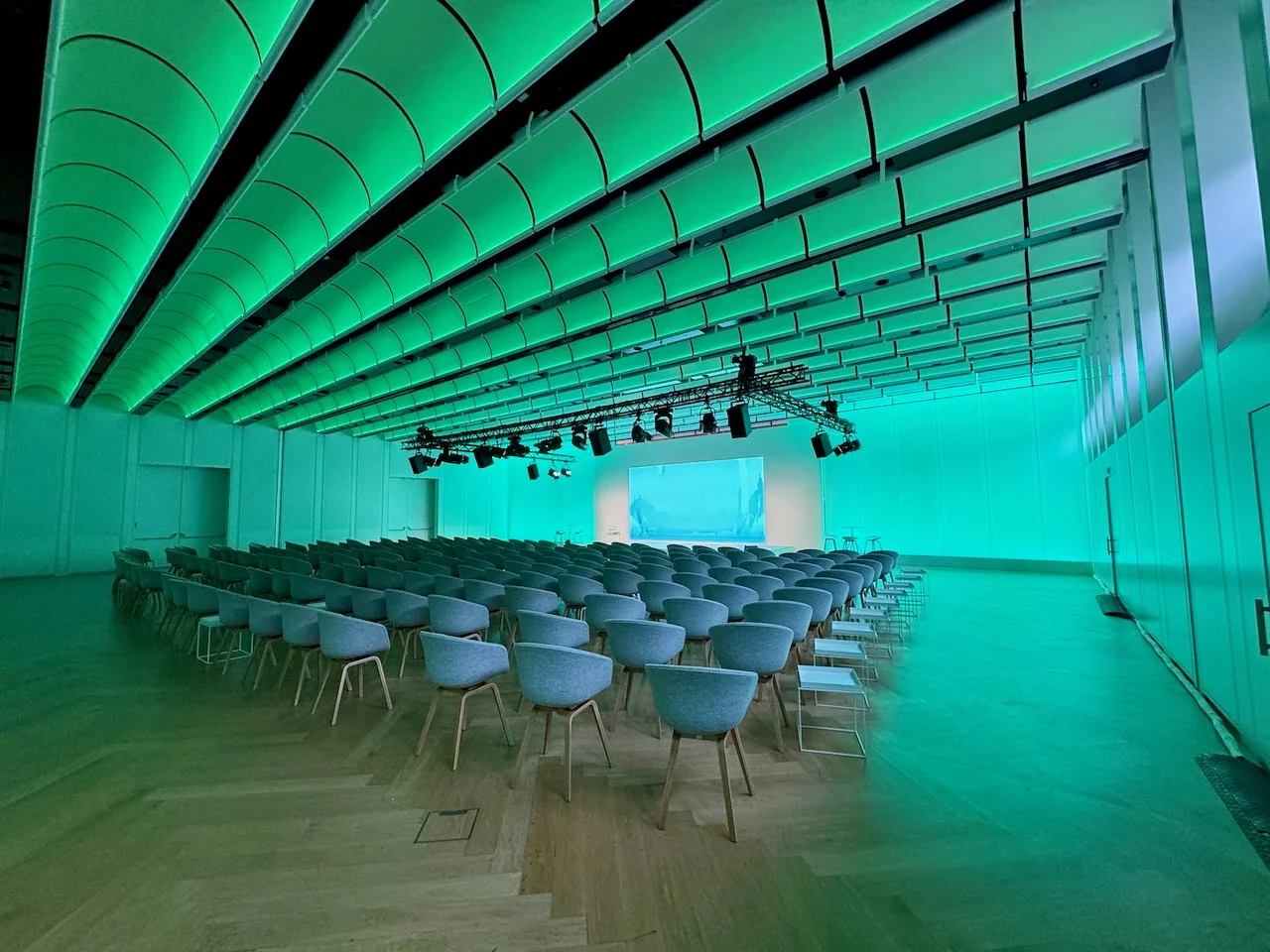 Conference room with rows of chairs facing a large screen, illuminated with green and blue lighting on the ceiling and walls. Minimalism is not easy.