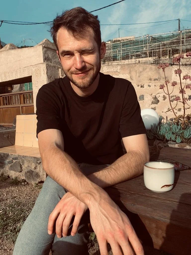 Ein junger Mann sitzt im Freien an einem Tisch, trägt ein schwarzes T-Shirt, und hat braune Haare und einen Bart. Der Himmel ist blau, und im Hintergrund sind Gebäudestrukturen, Gärten und eine Sukkulentenpflanze sichtbar.