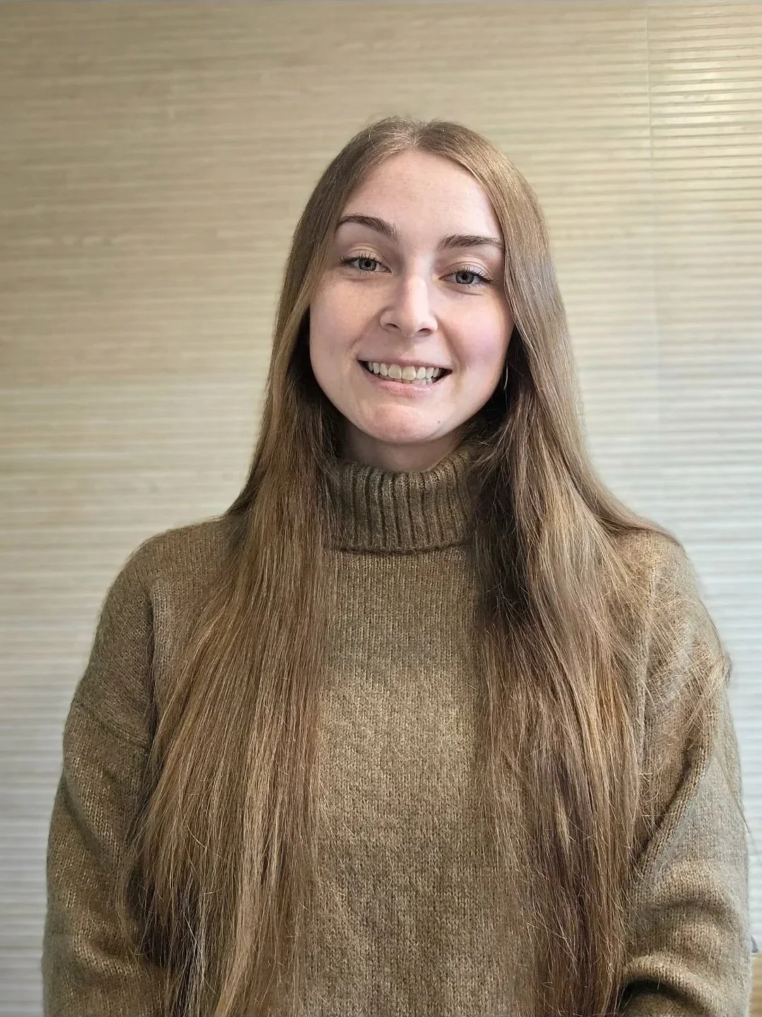 A young woman with long light brown hair, wearing a brown turtleneck sweater, smiling in front of a light beige textured background.