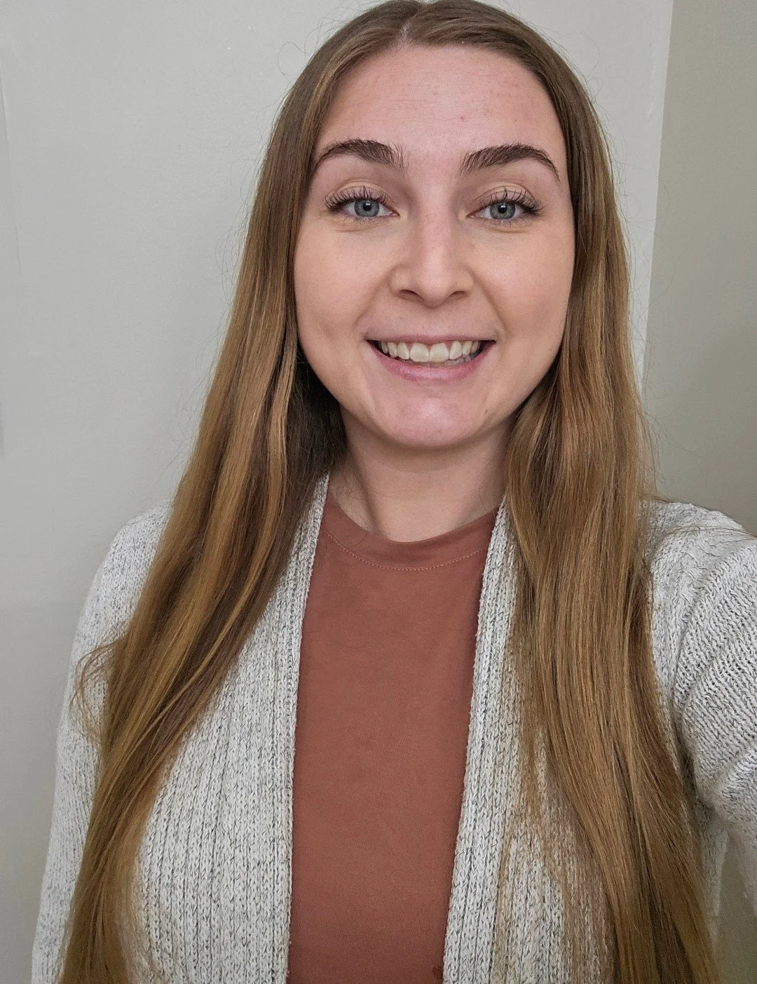 A woman with long red hair, blue eyes, and a bright smile, standing indoors against a plain light-colored wall, wearing a beige knit cardigan over a brown top.