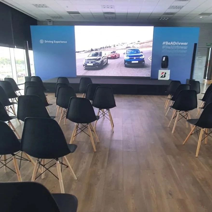 Sala de conferencias con sillas negras y una gran pantalla que muestra coches en un circuito de carreras, con logotipo de Volkswagen y hashtag #BeADriver.