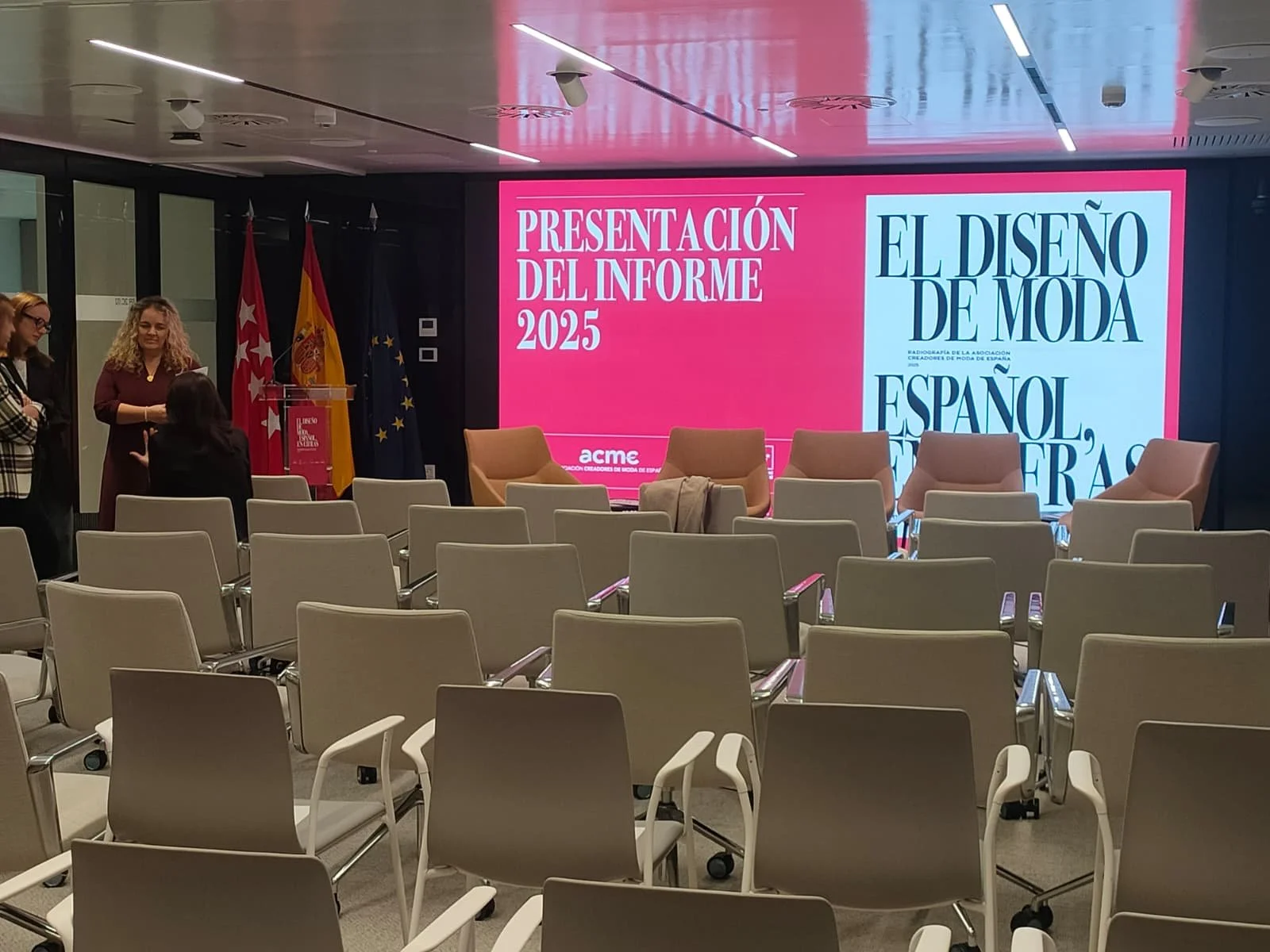 Sala de conferencias con sillas vacías, un escenario con pantalla grande que dice 'Presentación del informe 2025' y 'El diseño de moda español', y varias banderas en el fondo.