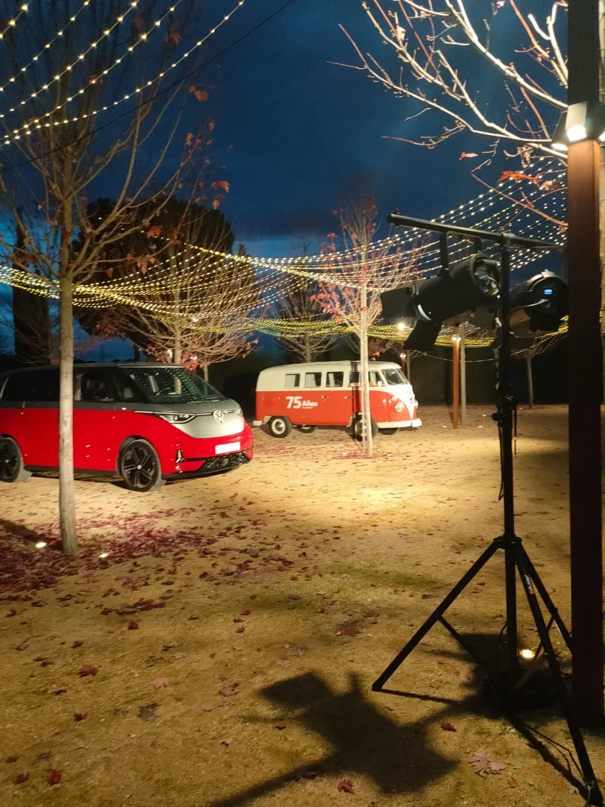 Escenario al aire libre con luces colgantes en los árboles durante la noche, con un coche y una camioneta de color rojo y blanco en el fondo. Hay focos en un soporte y hojas secas en el suelo.