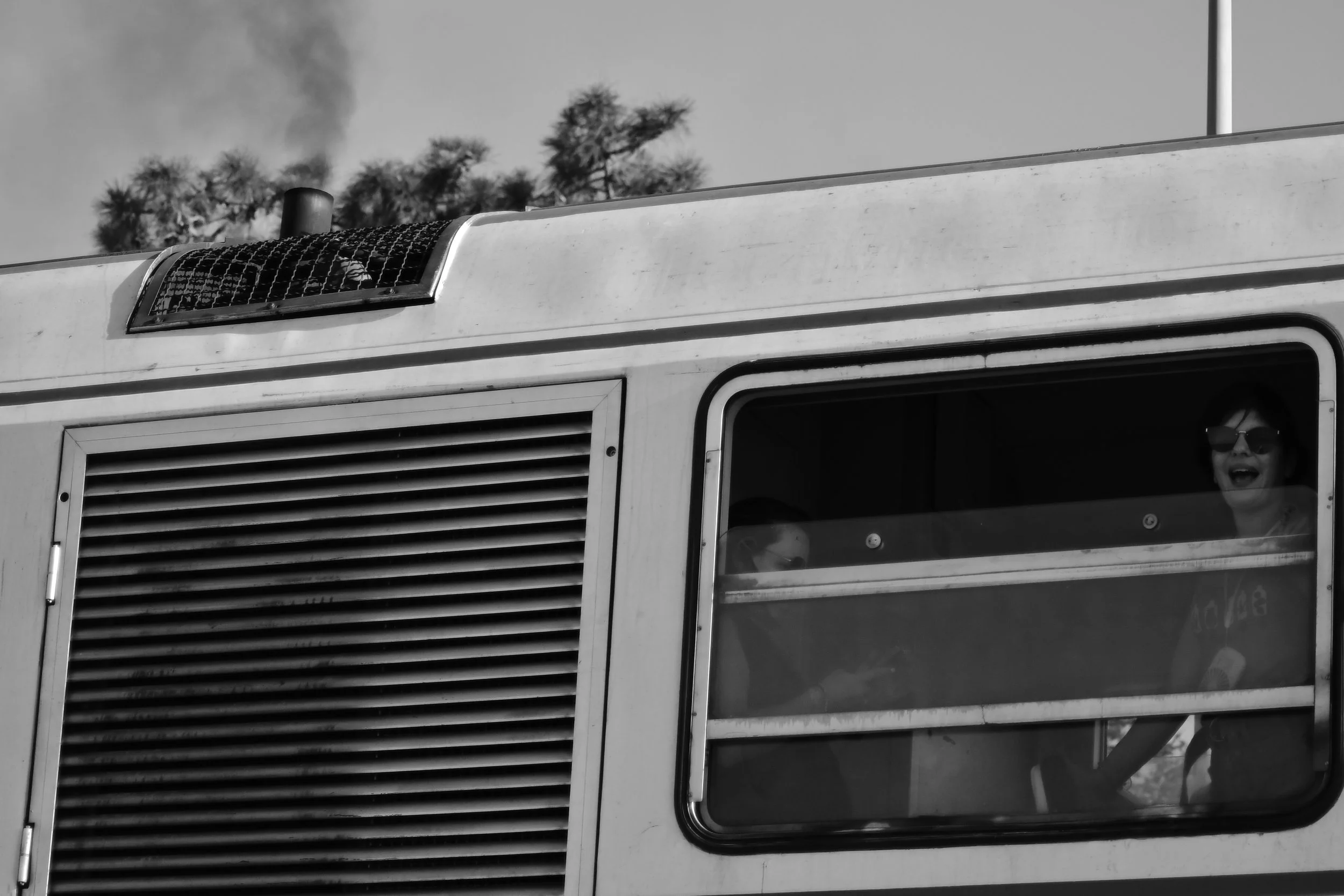 Photo en noir et blanc d'une partie d'un véhicule avec deux personnes visibles à travers une fenêtre. Une personne porte des lunettes de soleil et sourit.