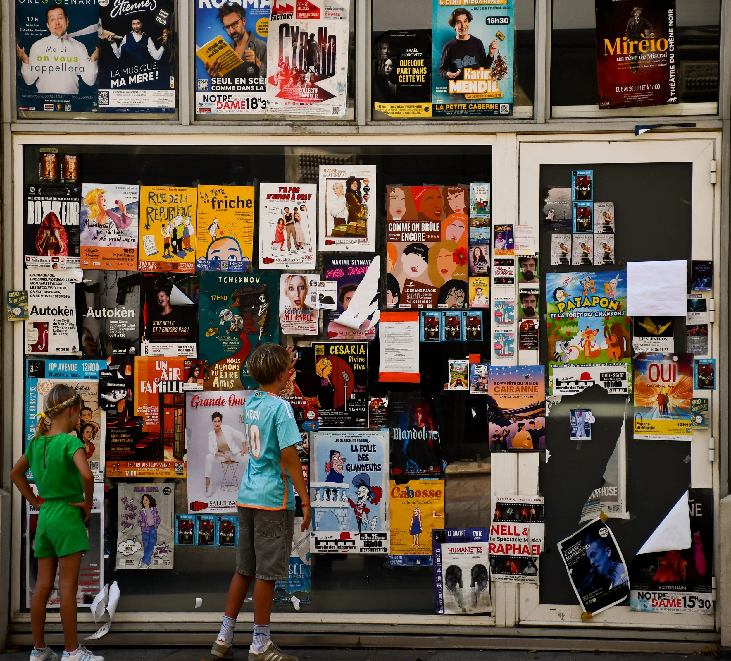 Une vitrine d'affiches de spectacles, de pièces de théâtre, de concerts et d'événements culturels, avec deux enfants observant l'affichage.