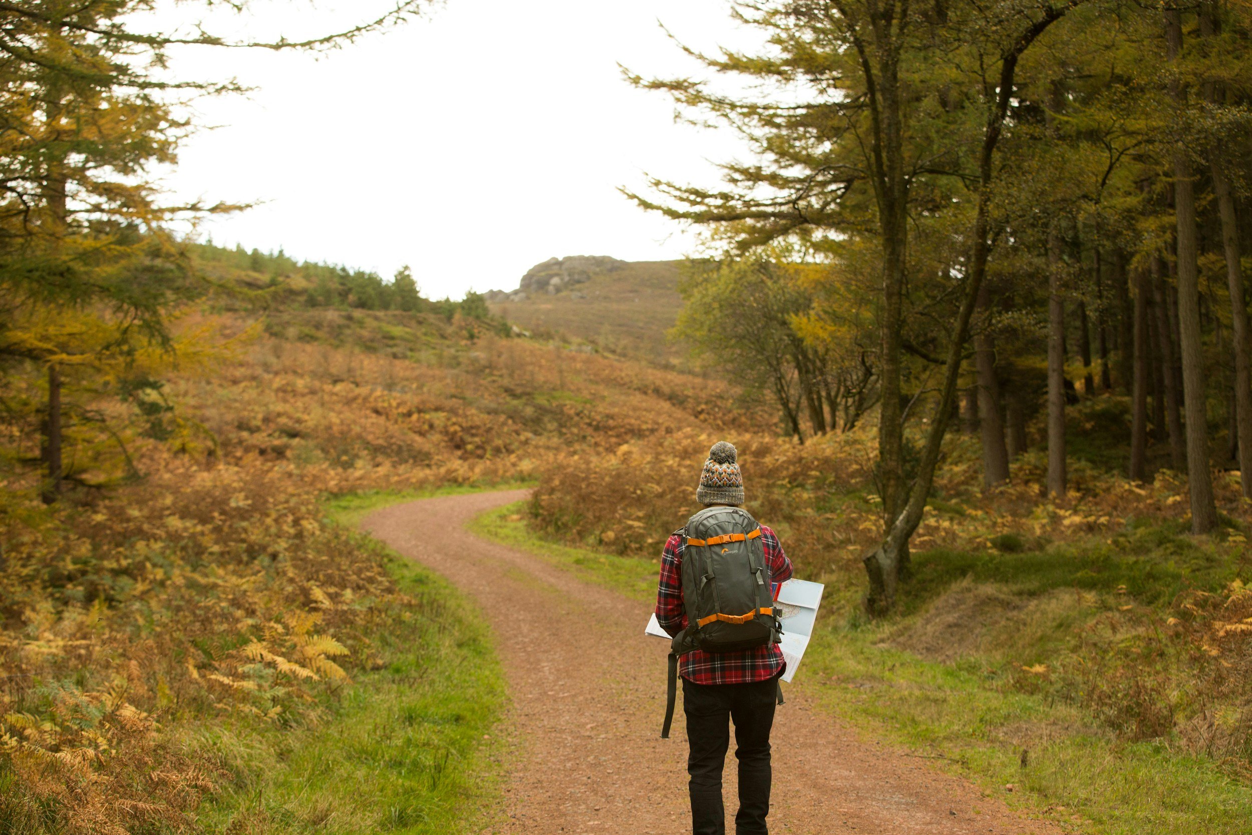 A person with a backpack and a beanie walking on a dirt trail through a forest with fall foliage.