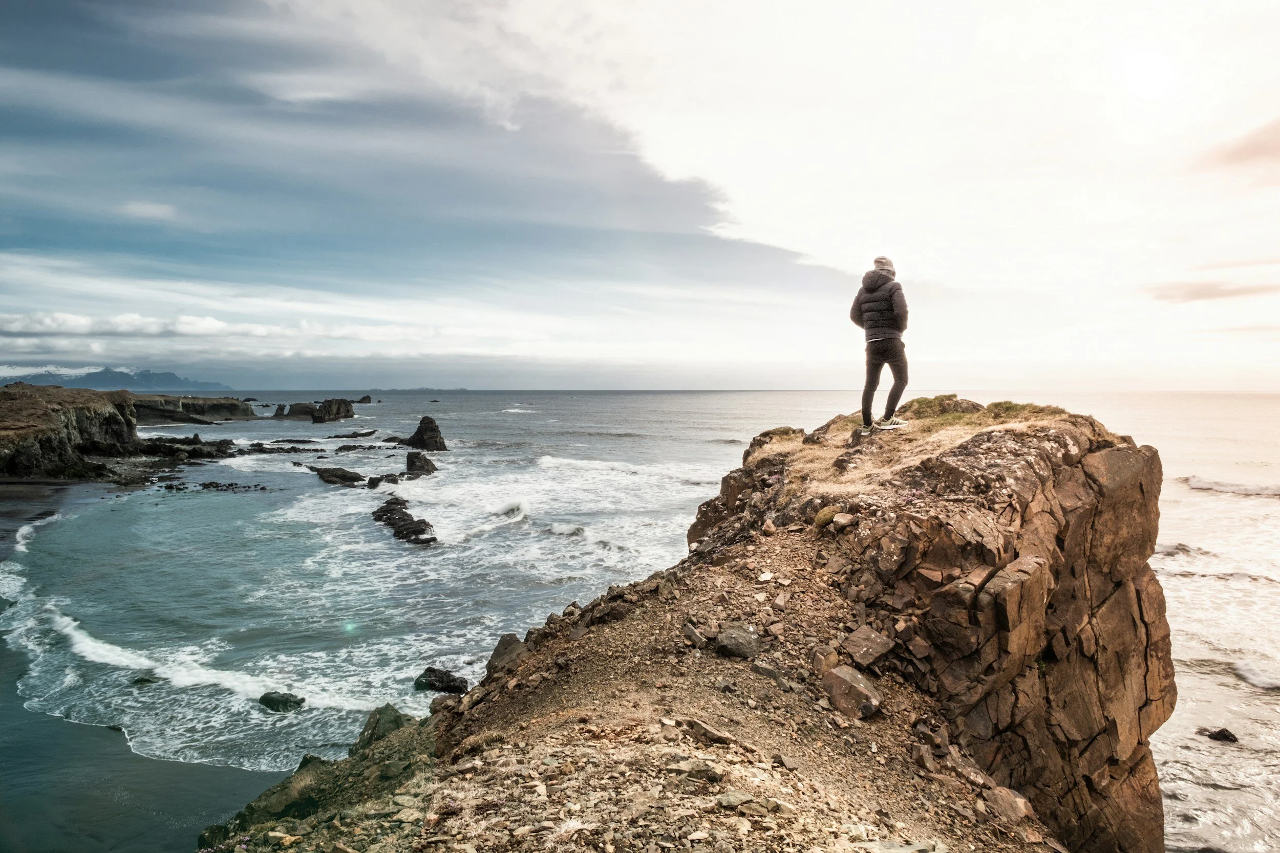 Person standing on a rocky outcrop overlooking the ocean during sunset or sunrise.