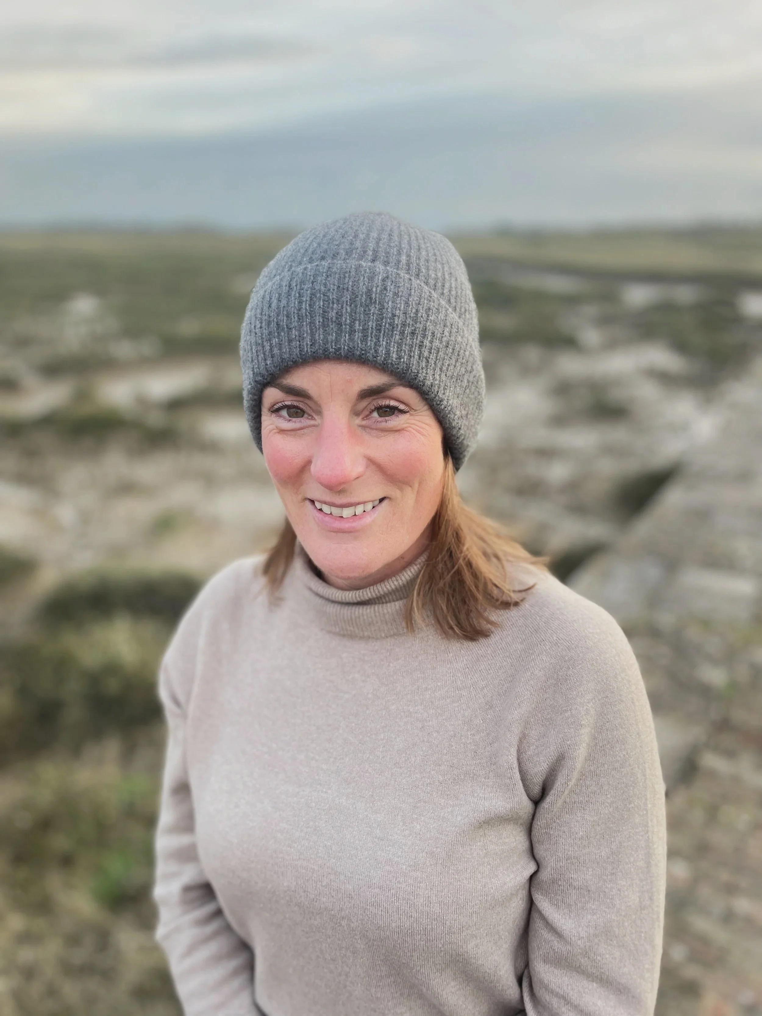 A woman with shoulder-length brown hair, smiling outdoors in a landscape by the sea with rocks and a cloudy sky.