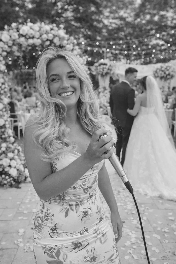 A woman holding a microphone smiles at a wedding reception, with a bride and groom dancing in the background under string lights and floral decorations.
