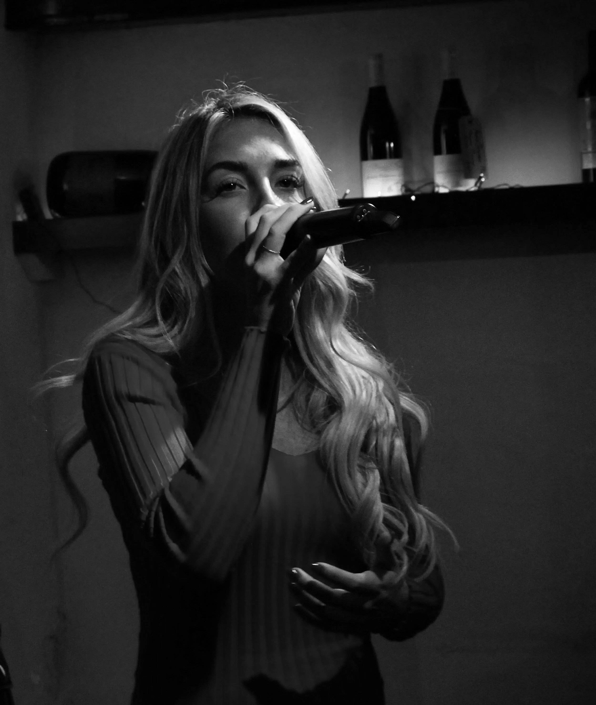 A young woman with long wavy hair holding a microphone close to her mouth, appearing to sing or speak, in a dimly lit room with bottles on a shelf in the background.