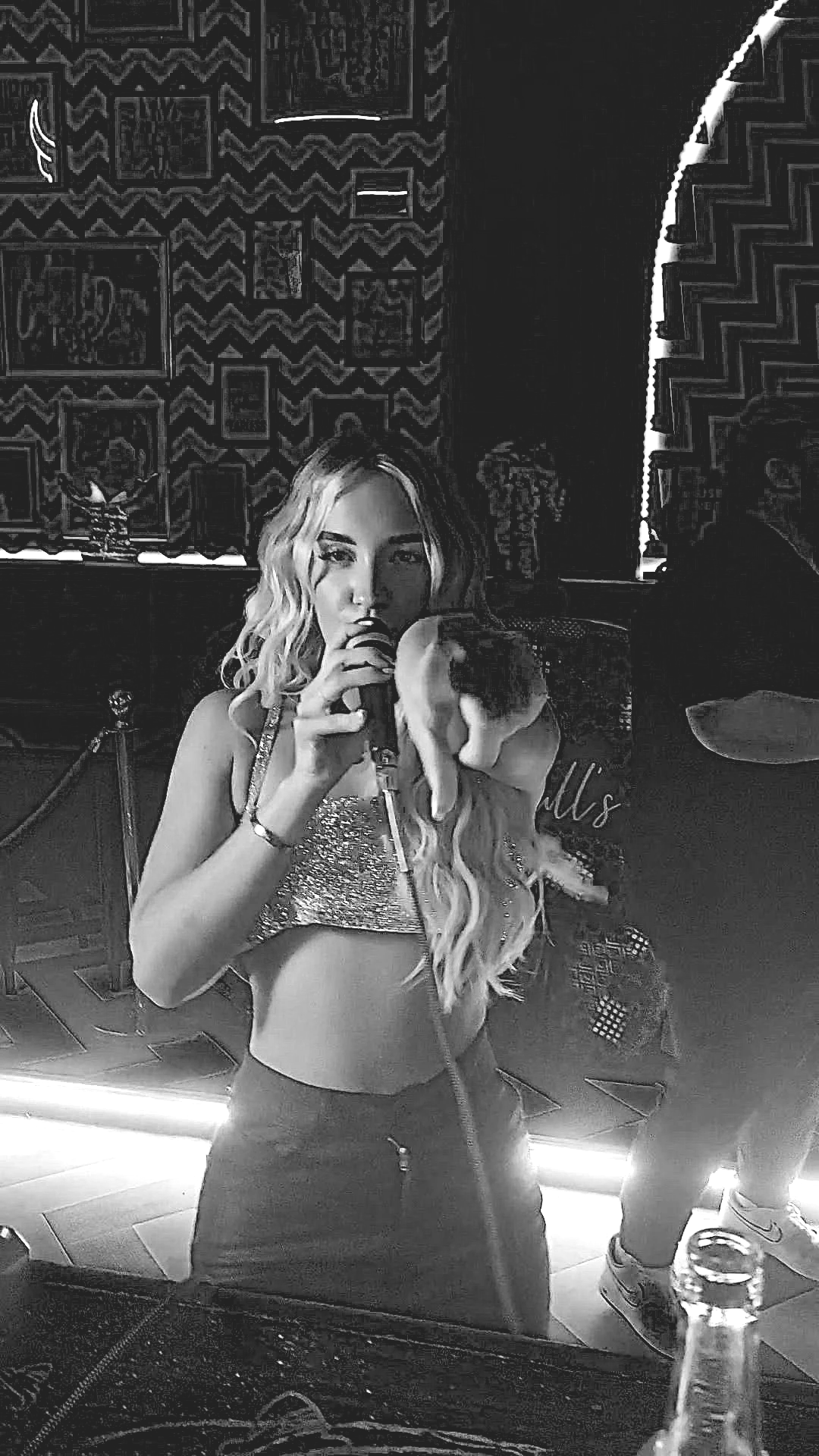 A woman with long wavy hair singing into a microphone in a dimly lit room with patterned wallpaper.